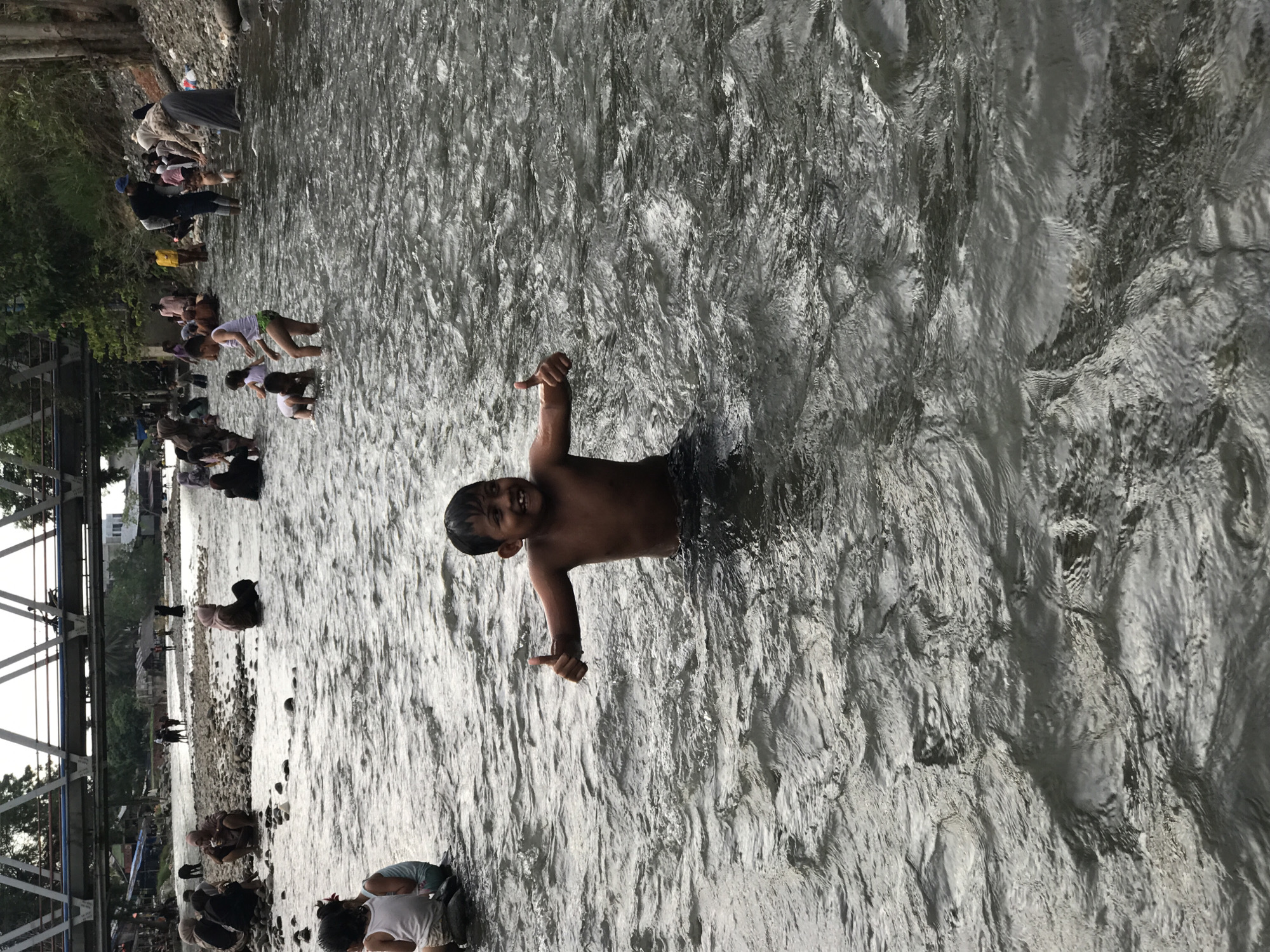 A boy is standing in a flowing river in Meurah, Indonesia. The river is filled with people bathing, washing, and standing in the water along the edges of the river. The river bank is to the left, and a bridge spans the river in the background. The boy is facing forward with his arms outstretched, giving a thumbs-up with each hand. He is smiling.