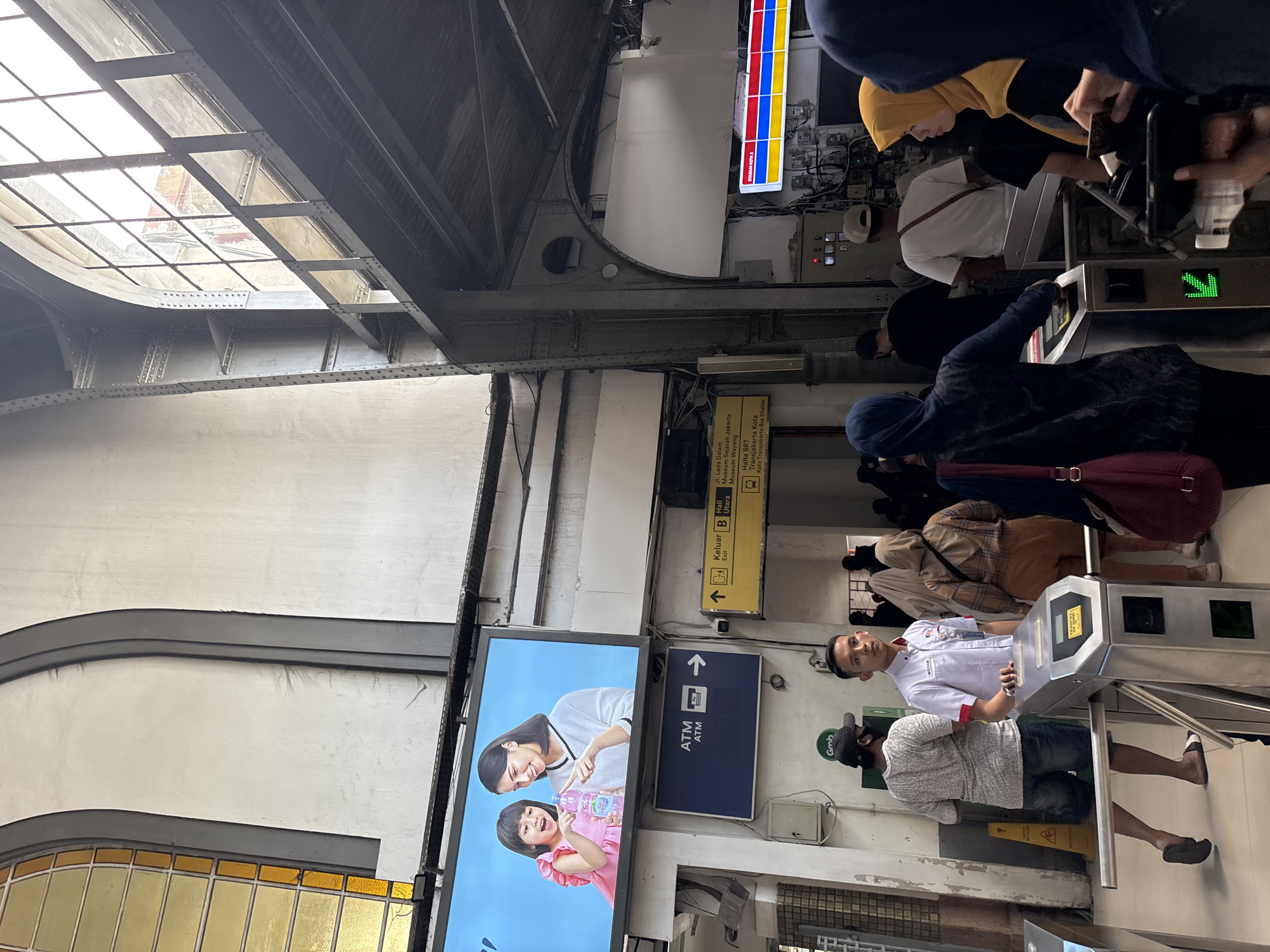The image captures an active public transit area, strongly identified as Kota Station (Stasiun Kota) in West Jakarta, Indonesia, given the numerous local signs. The scene showcases an architectural style featuring large, possibly arched, windows or skylights that allow natural light into the space, illuminating the sturdy, riveted metal beams and white-painted walls of the building. The bright lighting suggests it is daytime.
The primary activity involves several people passing through metal turnstiles, indicating entry or exit from a controlled access zone, likely a train or bus terminal. Visible subjects include a woman in a yellow hijab in the process of scanning something at a turnstile, and another person in a dark hoodie with a red bag also at a turnstile. Further back, a man in a white uniform shirt, possibly station staff, stands near the gates. In the foreground, another man wearing a patterned shirt, shorts, and a green cap with a "Grab" logo is also near a turnstile. The overall atmosphere appears routine, with commuters engaged in their daily transit.
Prominently displayed signage includes a large digital billboard featuring an advertisement with an adult woman and a child holding a water bottle, alongside a blue sign with an upward arrow indicating an "ATM." A key yellow directional sign, entirely in Indonesian, points to various local landmarks and facilities: "Keluar B" (Exit B), "Hall Utara" (North Hall), "Jl. Lada Dalam," "Museum Sejarah Jakarta," "Museum Wayang," "Halte BRT" (BRT Stop), "Transjakarta Kota," and "Kota Transjakarta Bus Shelter." A partially visible vertical sign in the upper right confirms the location as "STASIUN KOTA I."