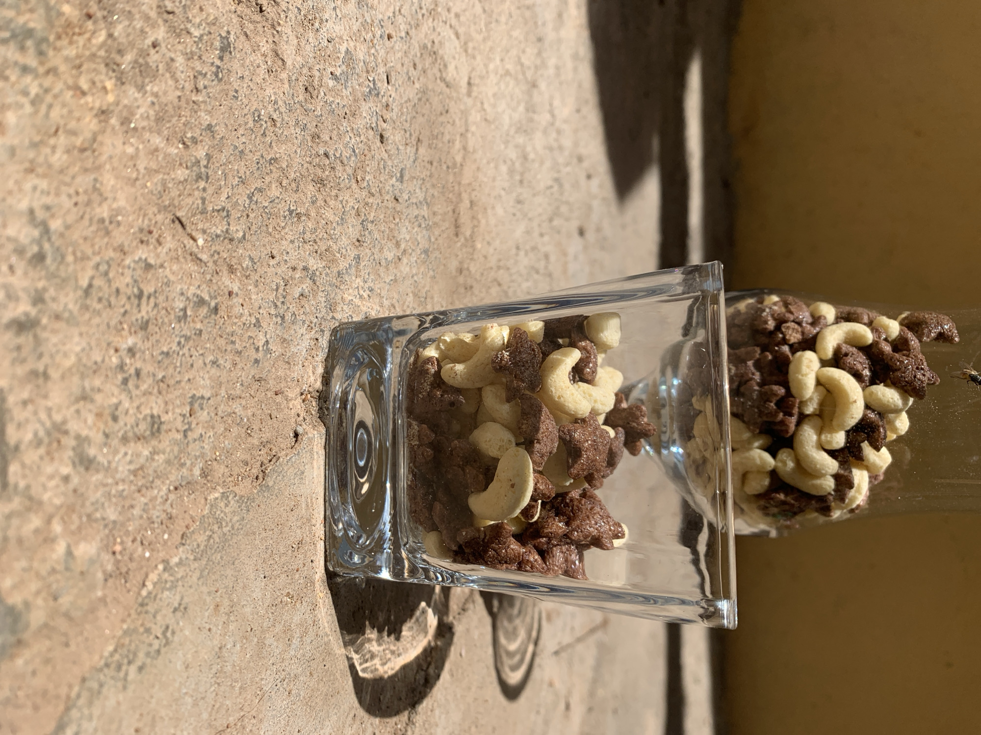 The scene, located in Rafin Par, Nigeria, depicts two transparent glass vessels containing dry cereal, positioned on a coarse, light brown concrete substrate. The foreground vessel is a square-bottomed glass, partially filled with a mixture of light-colored, cashew-shaped cereal pieces and dark brown, star-shaped cereal pieces. The base of this vessel exhibits concentric circular indentations. The second vessel, situated behind and above the first, is also transparent glass and contains an identical cereal mix. A small, dark arthropod is visible in the upper left region of the image, proximate to the rim of the rear vessel. The background comprises an indistinct, solid, light brown-yellow wall. Direct illumination, presumably sunlight, originates from the right, producing sharply defined shadows of the vessels and their contents on the concrete surface and the wall. No human figures or overt actions are present.