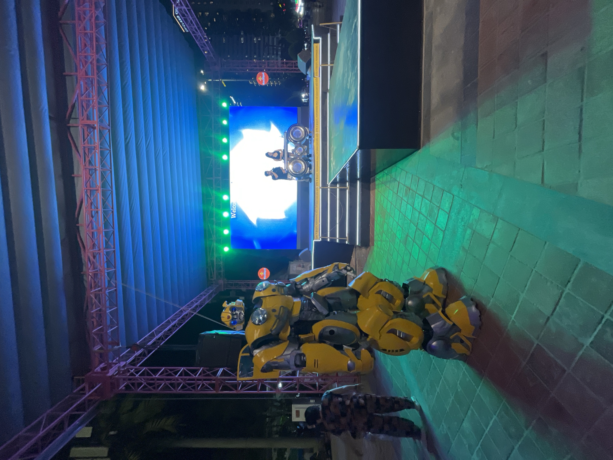 This image captures an outdoor event stage setup at night, possibly in Đà Nẵng, Vietnam. The primary focus is a large, detailed replica of the character Bumblebee from the Transformers franchise, positioned in the foreground. The robot is predominantly yellow with gray accents and appears to be in a dynamic pose, suggesting it might be part of a performance or display.
Behind the robot, a stage is set up with a large LED screen displaying a bright white arrow pointing to the right, with the word "Welcome" visible in the bottom left corner of the screen. Three individuals, dressed in dark clothing, are visible on the stage in front of the screen, appearing to be performers or speakers. The stage is lit with green and blue lights, creating a vibrant atmosphere.
The scene is framed by industrial-looking pink scaffolding and a large, pleated blue backdrop. To the right of the stage, there's a raised platform with green tiled flooring, illuminated by green lighting that casts a distinct glow.
Below the stage and in front of the Bumblebee replica, two individuals in camouflage clothing are visible, walking away from the camera. Their presence suggests they might be event staff or security.
The overall setting indicates an entertainment event, likely a concert, festival, or product launch, taking place after dark. The combination of lighting, the prominent robot prop, and the stage setup suggests a lively and engaging atmosphere. There is no clear indication of weather conditions, but the nighttime setting is evident from the artificial lighting and the dark sky.