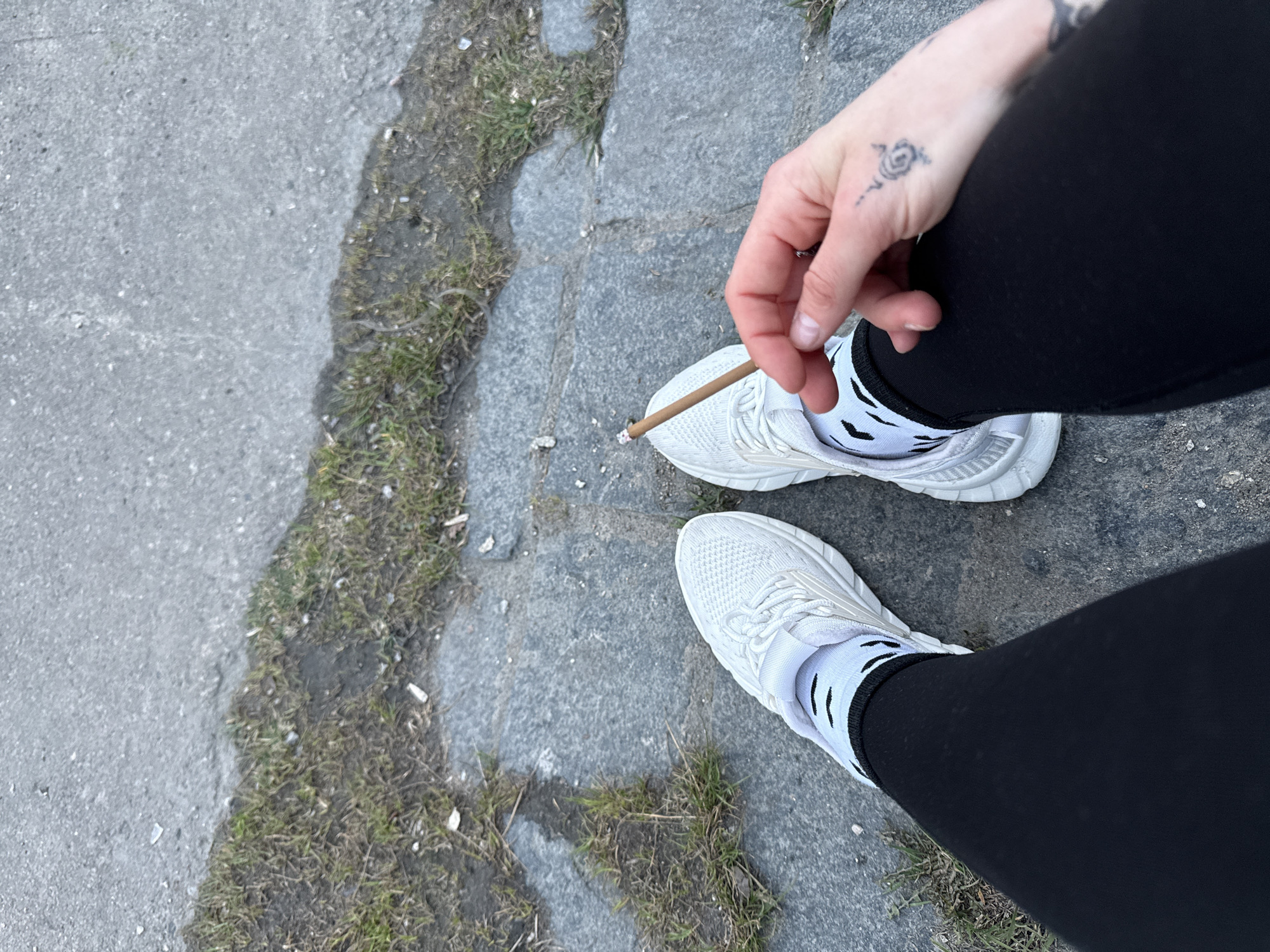 This high-angle, close-up image captures the lower body of a person standing or sitting outdoors in Saint Petersburg, Russia. The perspective looks downwards, showing the person's feet, legs, and one hand.
The individual is wearing black leggings or pants, paired with white low-cut socks featuring black, abstract markings. On their feet are chunky white athletic sneakers with visible lacing and textured soles. The person's right hand, adorned with a small, stylized rose or spiral tattoo on its back, is holding a lit, hand-rolled cigarette or joint, identifiable by the ember at its tip.
The ground is a mix of urban surfaces: a lighter grey asphalt or concrete area on the left transitions into darker, irregularly shaped grey cobblestones or stone slabs towards the center and right. Patches of dirt and sparse, dry green and brown grass grow in the crevices between the stones and along the edges of the paved areas. The lighting suggests it is daytime, with no strong shadows indicating direct sunlight, possibly an overcast day. No specific emotions are discernible, and no text is visible in the frame. The scene depicts a casual, private moment of smoking outdoors.