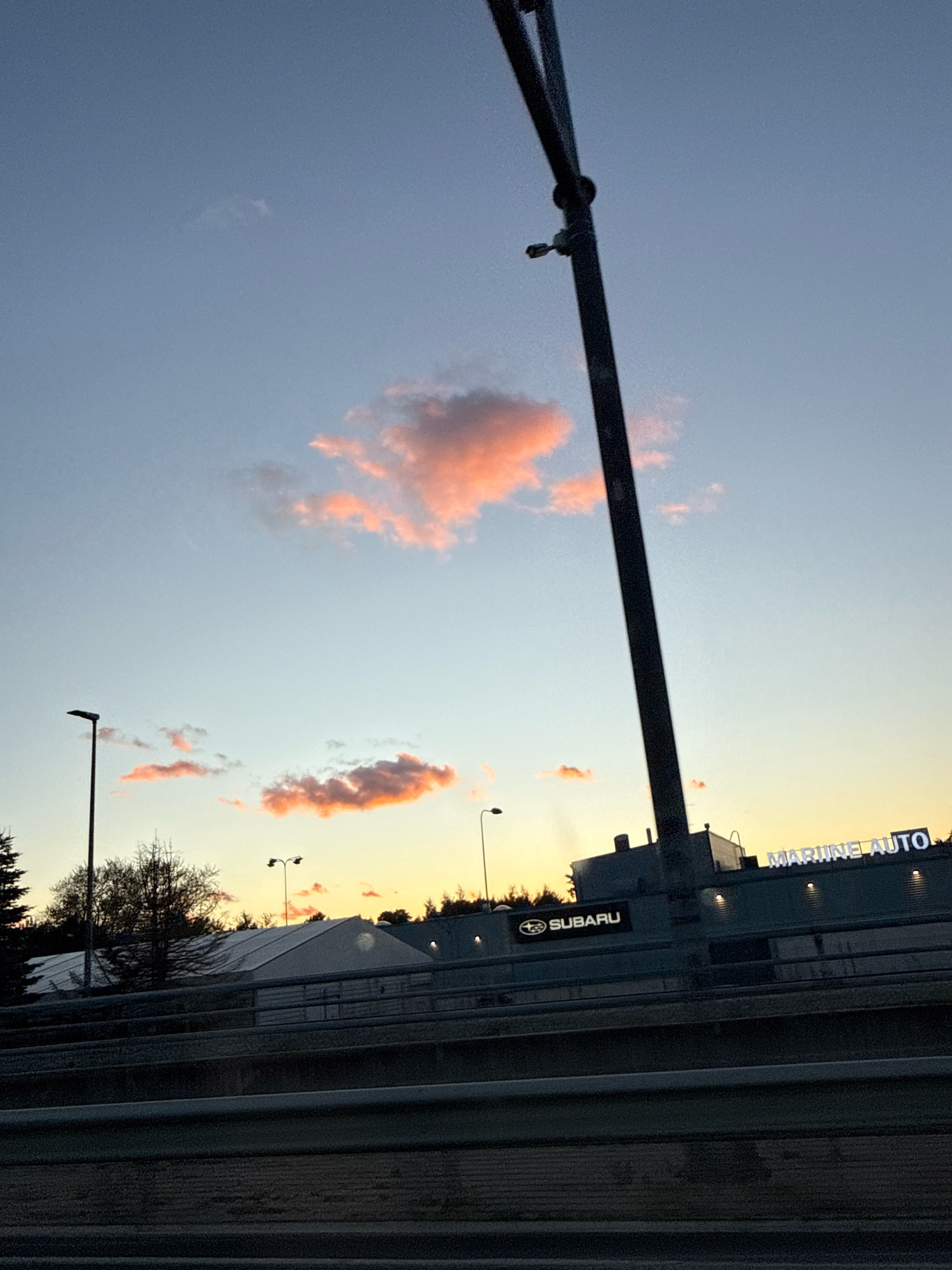The image depicts a street view in Tallinn, Estonia, at dusk. The sky is a gradient of pale blue to orange, with a few wispy pink clouds. A tall, dark, diagonal pole dominates the right side of the frame, extending from the top edge downwards. In the middle ground, a modern-looking building with lit signage that reads "MARIINE AUTO" is visible. To the left of this building, a black rectangular sign with the Subaru logo is also illuminated. Behind the buildings, a line of trees forms a dark silhouette against the colorful sky. In the foreground, a dark, ribbed barrier or railing runs horizontally across the bottom of the image. Streetlights are visible in the middle ground and background, and a large, white tent-like structure is also present behind the barrier. The overall impression is a commercial area during twilight.