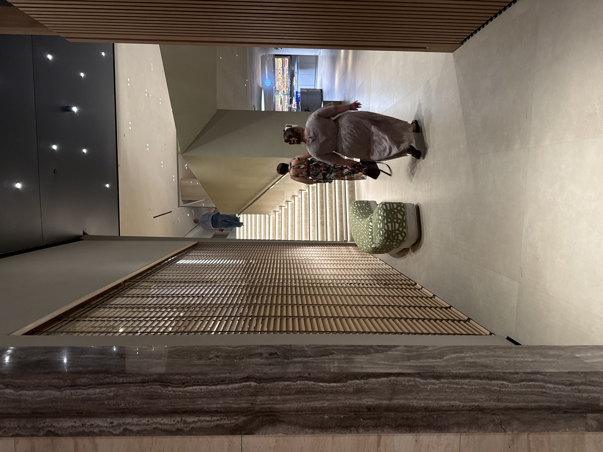 The image depicts a contemporary interior space with a light-colored tiled floor. In the foreground, a person in a light brown, floor-length dress walks away from the camera, carrying a dark handbag. To the left, a patterned green bench with a curved design is visible. Further into the scene, a staircase ascends to the right, with a person in a blue shirt and dark pants walking up it. Two women in floral dresses are also seen walking up the stairs, one ahead of the other. The environment features modern architecture with a large wall covered in wooden blinds on the left, and a wall with vertical wooden slats on the right. In the background, a retail or dining area is visible with displays and lighting. The overall impression is an indoor public space with an architectural focus on materials and modern design. The location is specified as Симеизкий поселковый совет, Ukraine.