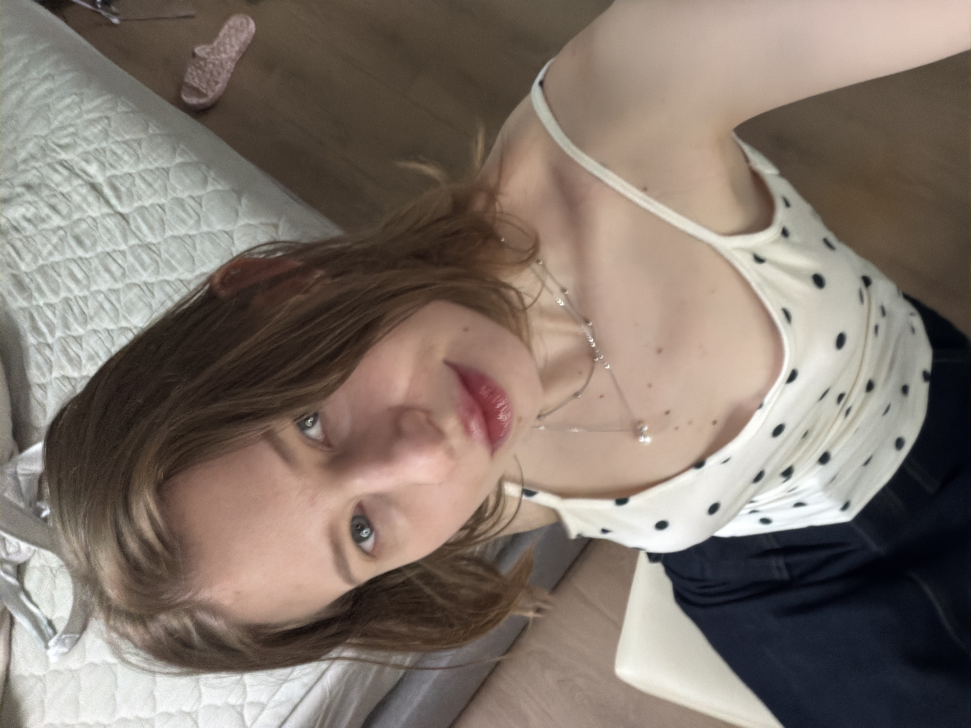 A young Caucasian woman with shoulder-length brown hair and blue eyes is looking up at the camera. She is wearing a white tank top with black polka dots and a delicate silver necklace with a pearl pendant. Her lips are painted a glossy red. She is sitting on a white chair, and her dark blue jeans are visible. In the background, a bed with a textured, light-colored comforter is visible, along with a pink slipper on the wooden floor. The lighting suggests an indoor setting, likely a bedroom. The image appears to be a selfie, taken from a high angle.