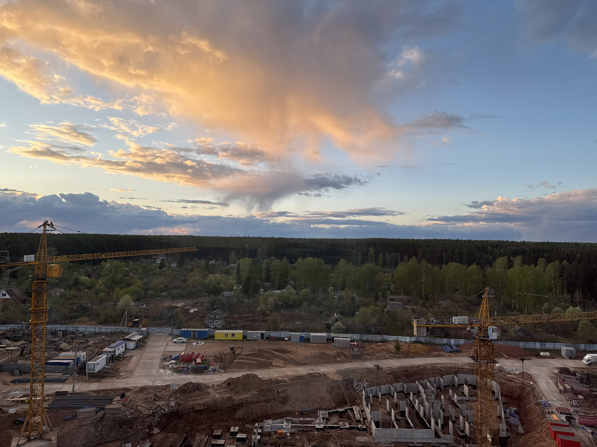 The image shows a construction site in Izhevsk, Russia, taken during the late afternoon or early evening. The sky is partially cloudy with a mixture of white, orange, and gray clouds. A band of green trees is present in the midground, behind what appears to be a semi-urban area with scattered buildings.
In the foreground, a construction site features a concrete foundation with several vertical concrete pillars. Yellow cranes are visible on the left and right sides of the frame. Construction materials and temporary structures are scattered throughout the construction area.
