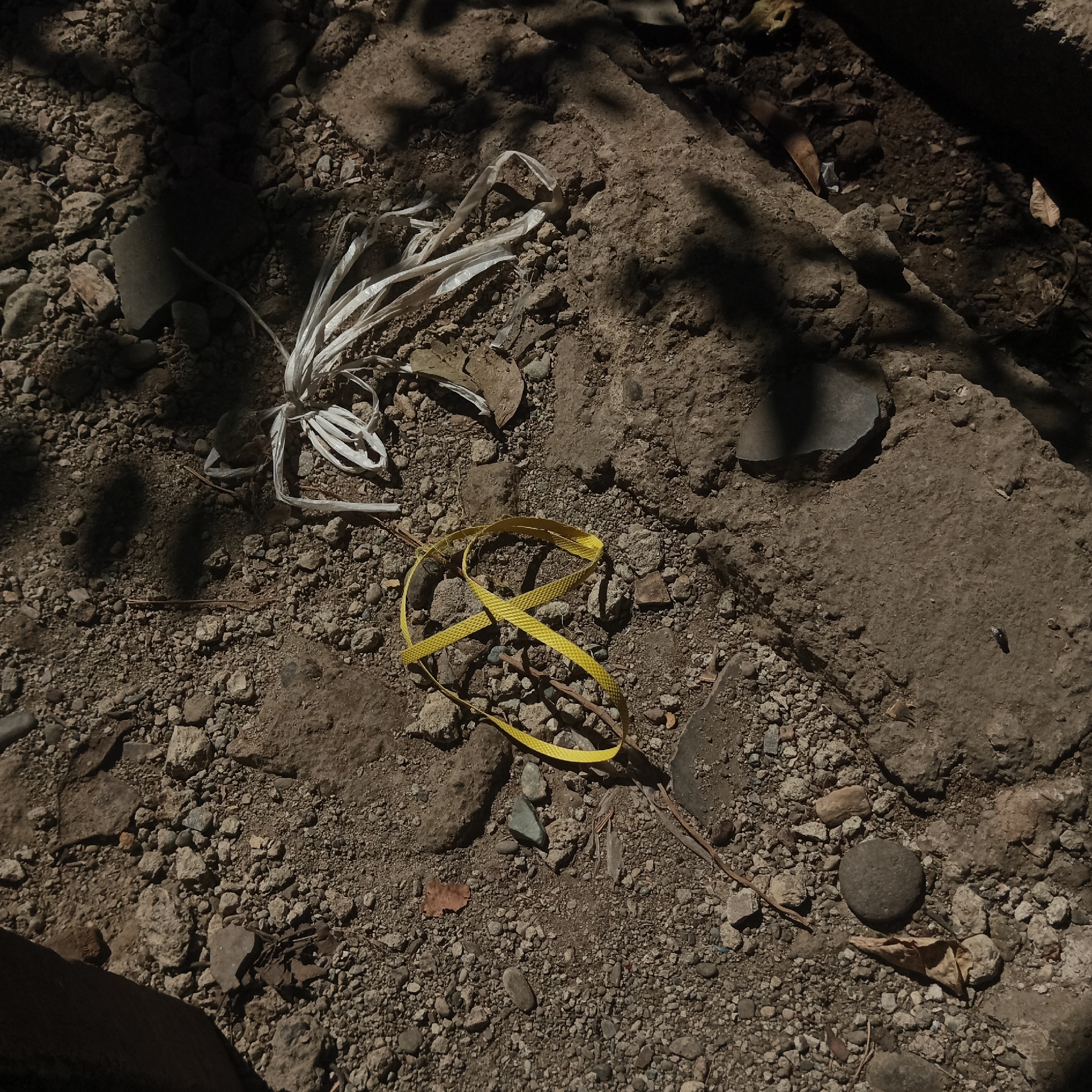 The image depicts a close-up, overhead view of a gritty, rocky ground surface, likely a path or unpaved area in Tumampua, Indonesia. Sunlight casts dappled shadows from overhead foliage onto the earth, highlighting the textures of loose stones, dirt, and debris.

Prominently featured are two pieces of discarded man-made material. In the upper left quadrant, a tangled mass of white plastic strapping lies on the ground. To its right and slightly lower, a bright yellow measuring tape is coiled in a loop, its reflective surface catching the light. The surrounding ground is a mixture of small pebbles, larger rocks, and dark soil. Scattered among the rocks are a few dry leaves and thin twigs.

The overall impression is one of a natural environment interrupted by human-generated waste. The lighting and composition emphasize the textural details of the ground and the contrast between natural and artificial elements.