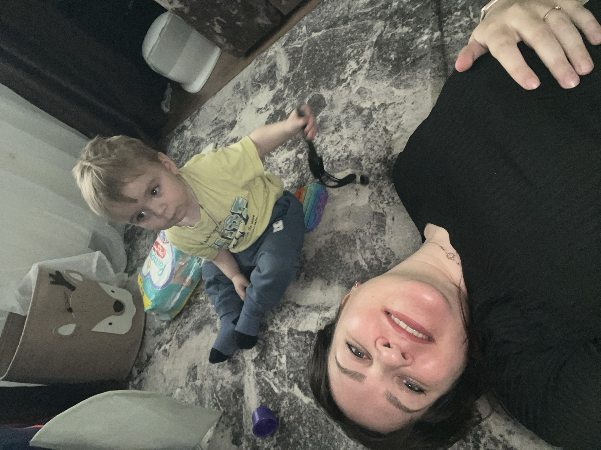 A woman lies on a gray rug next to a young child in a yellow shirt and blue pants, who is sitting and holding a black object. The rug is in a room with a beige container with a deer face, a package of diapers, and other items. The woman is smiling, and her face is close to the camera. A ring is visible on her finger and a bracelet on her wrist. The location of Noviy, Russia is not discernable in the image.