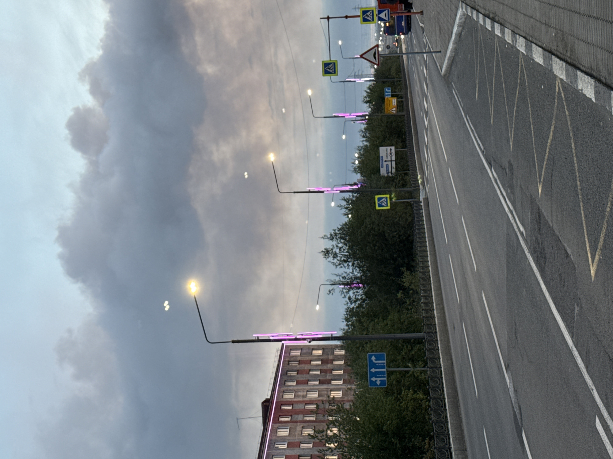 The image depicts a street scene in Norilsk, Russia, under a cloudy sky. A multi-lane asphalt road runs through the frame, marked with white lane dividers. On the left side of the road, a sidewalk is bordered by a metal fence and lush green trees. A tall streetlamp with a bright yellow light stands prominently, and another streetlamp with purple neon accents is visible. Further down the street, more streetlights are present, some with purple lighting.
A large, multi-story building with a patterned facade and illuminated windows is situated behind the trees on the left. Numerous road signs are scattered along the roadside, including pedestrian crossing signs with a yellow background and a silhouette of a person, a yield sign, and directional signs.
In the distance on the right side of the road, a bus is visible, along with another truck. The area is characterized by urban infrastructure, including streetlights, signs, and buildings, suggesting a typical city environment. The overall atmosphere is subdued due to the cloudy weather and the twilight hour.