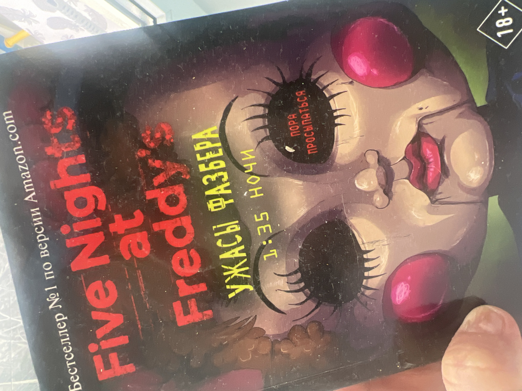 A close-up shot depicts a book cover titled "Five Nights at Freddy's" with the subtitle "Fazbear's Horrors" in Russian. The cover features a creepy doll-like face with dark, empty eyes and bright red cheeks. Text on the cover reads "1:35 AM" and "Time to wake up" in Russian. The top right corner of the book indicates an 18+ age restriction. The book cover is held by a human hand. The background appears to be a room interior. The location is not specified, although the book cover is written in Russian. There is no evidence indicating that the image was taken in the city of Chaikovsky, Russia.