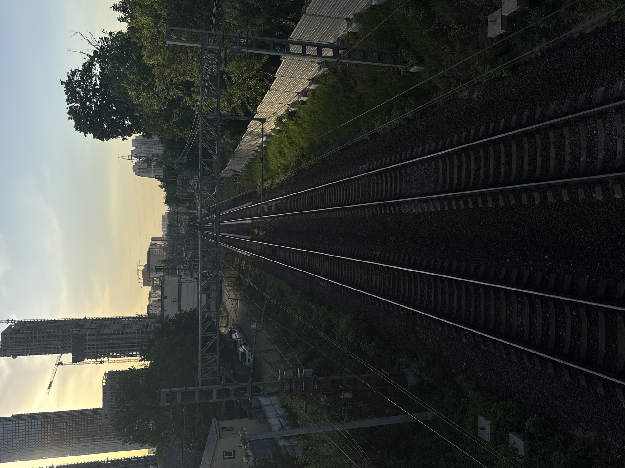 The image depicts a railway track stretching into the distance in Khimki, Russia. The track is flanked by vegetation on both sides. Overhead power lines and supporting structures run parallel to the rails. In the background, buildings, including those under construction with cranes, are visible against a sky. A noise barrier is present along one side of the track. A few vehicles are seen on the roadside.