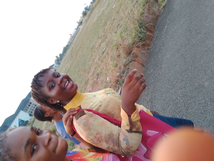 Three young women are standing on a gray asphalt road in Umueshi, Nigeria. The women are smiling and appear to be taking a selfie. Behind them is a grassy field. In the distance, there are buildings and a clear sky.