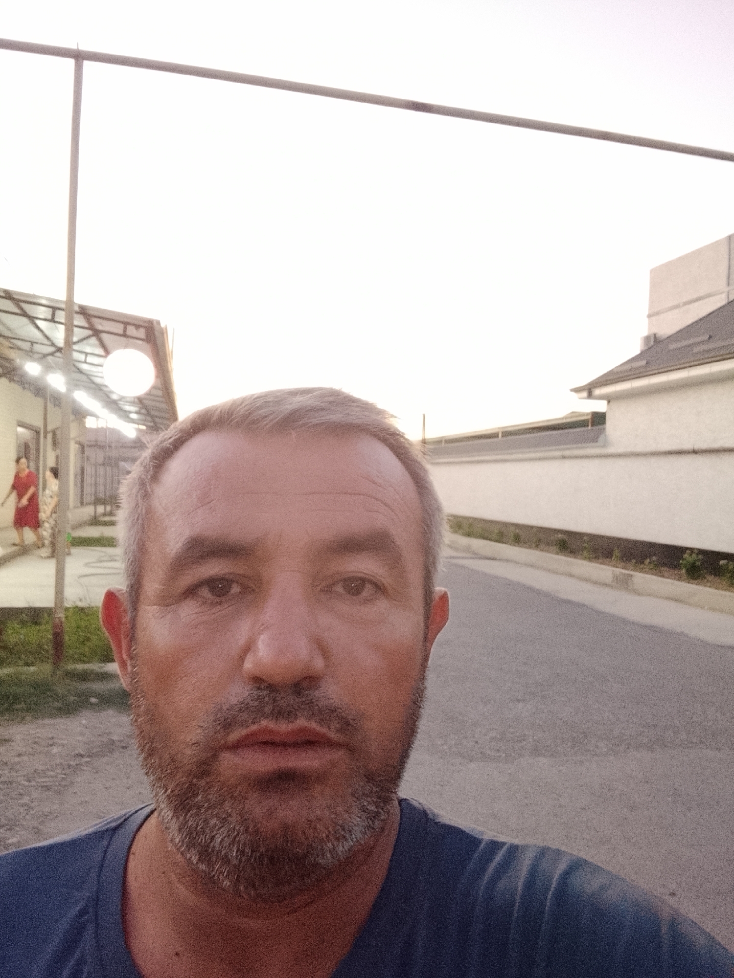 A mid-shot, selfie-style photograph shows a man with short, graying hair and a short beard. He is wearing a dark blue t-shirt. He is standing outdoors in front of a building with a light-colored facade and a dark-tiled roof. Behind him, to the left, a woman in a red dress is walking. A paved road is visible in the background. The sky is overcast and bright. The overall scene appears to be a residential or commercial area in Sabir Rakhimov, Uzbekistan, likely during the daytime.