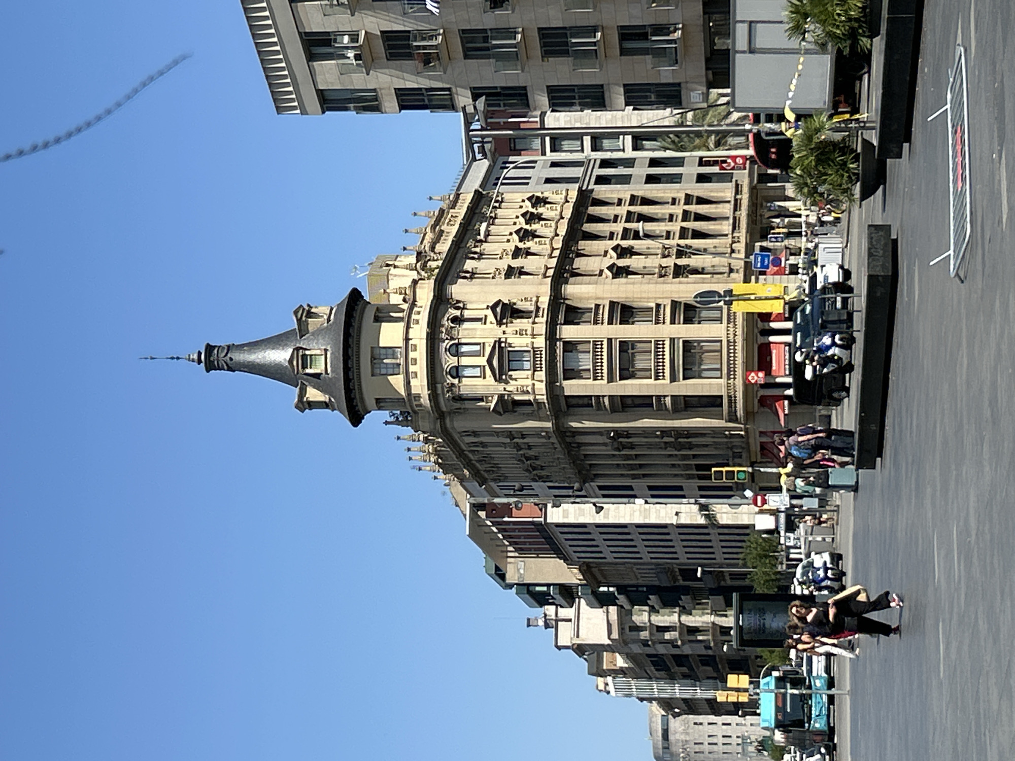 The image depicts a street scene in Barcelona, Spain, under a clear blue sky. In the foreground, a wide, paved intersection with a pedestrian crossing is visible. Several people are walking across the intersection, including a woman in black pants and a black top carrying a bag. A blue bus and several cars are also present on the street.
Dominating the background is a distinctive building with a conical, dark-colored turret, featuring multiple windows and ornate architectural details. This building is flanked by other multistory buildings of varying architectural styles, some with modern glass balconies. To the right, a large white truck and planters with palm trees are visible. The overall impression is that of a bustling urban environment on a sunny day.