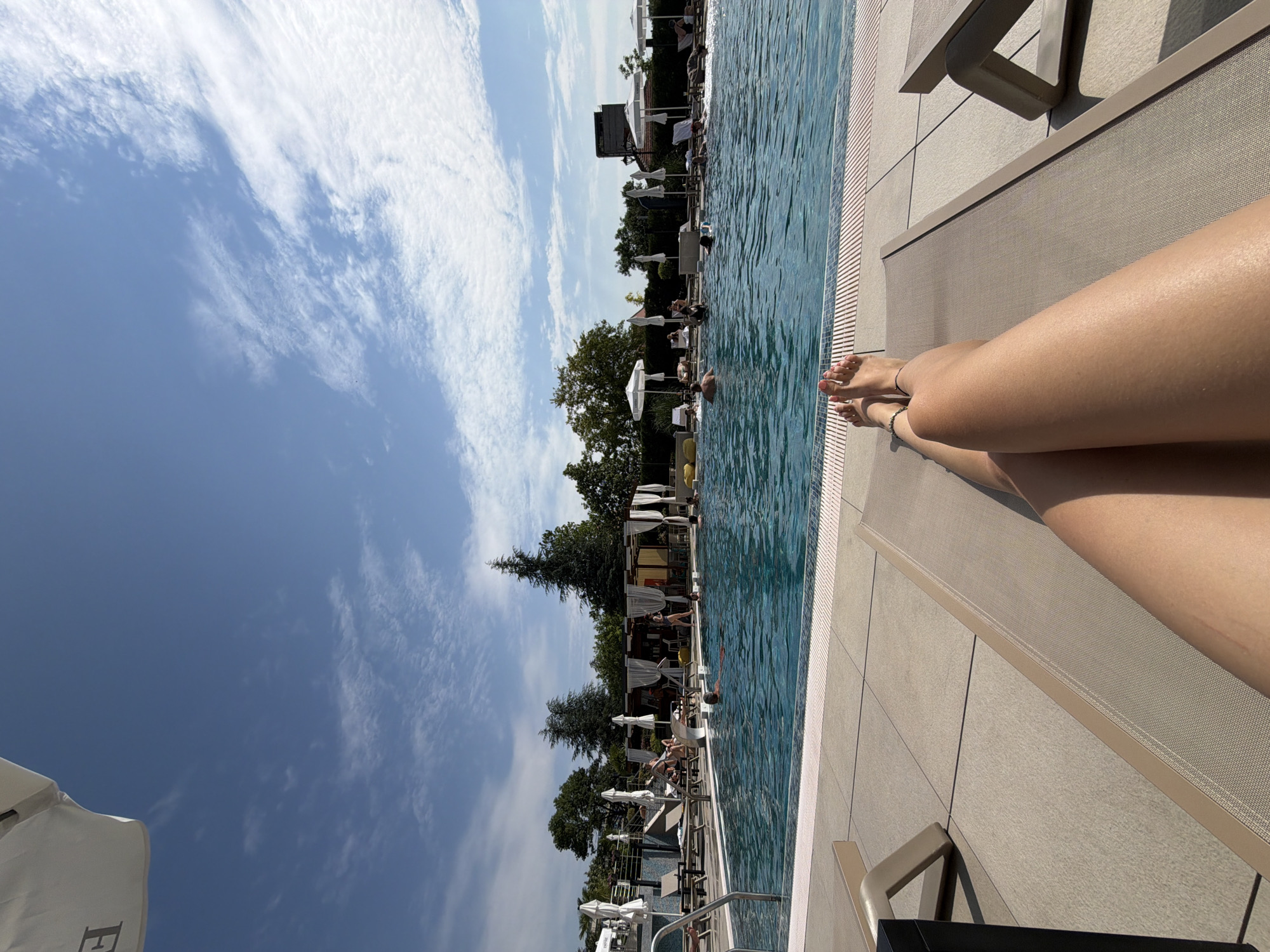The image depicts a sun-drenched scene at a swimming pool, likely located in Hisarya, Bulgaria, given the context provided. In the foreground, a person's legs are stretched out on a reclined lounge chair, their feet visible with toes pointed upwards. The person is wearing a delicate ankle bracelet on their left ankle. Beyond the lounge chair, a large, clear blue swimming pool dominates the mid-ground. Several people are visible in the pool, some swimming and others standing. The poolside area is paved with light-colored tiles and lined with lounge chairs and umbrellas, suggesting a resort or hotel setting. In the background, lush green trees form a backdrop, and a covered structure with white curtains and yellow seating is visible near the pool's edge, possibly a bar or dining area. The sky above is a brilliant blue, dotted with scattered white clouds, indicating a clear and pleasant day. The overall atmosphere is one of relaxation and leisure.