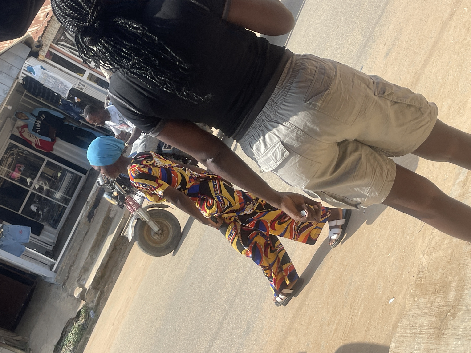 In Abuja, Nigeria, a bright, sunny day illuminates a street scene where two women are walking. The woman in the foreground, facing away from the camera, wears a black t-shirt and light-colored cargo shorts. Her braided hair is long and dark. The second woman, to her left and slightly ahead, is dressed in a vibrant, multi-colored patterned outfit, including a headscarf and wide-legged pants. She is wearing white sandals. Behind them, a motorcycle is parked, and further in the background, a shop displays clothing on racks outside its entrance. The street itself is paved with asphalt, and the surrounding environment suggests an urban setting.
