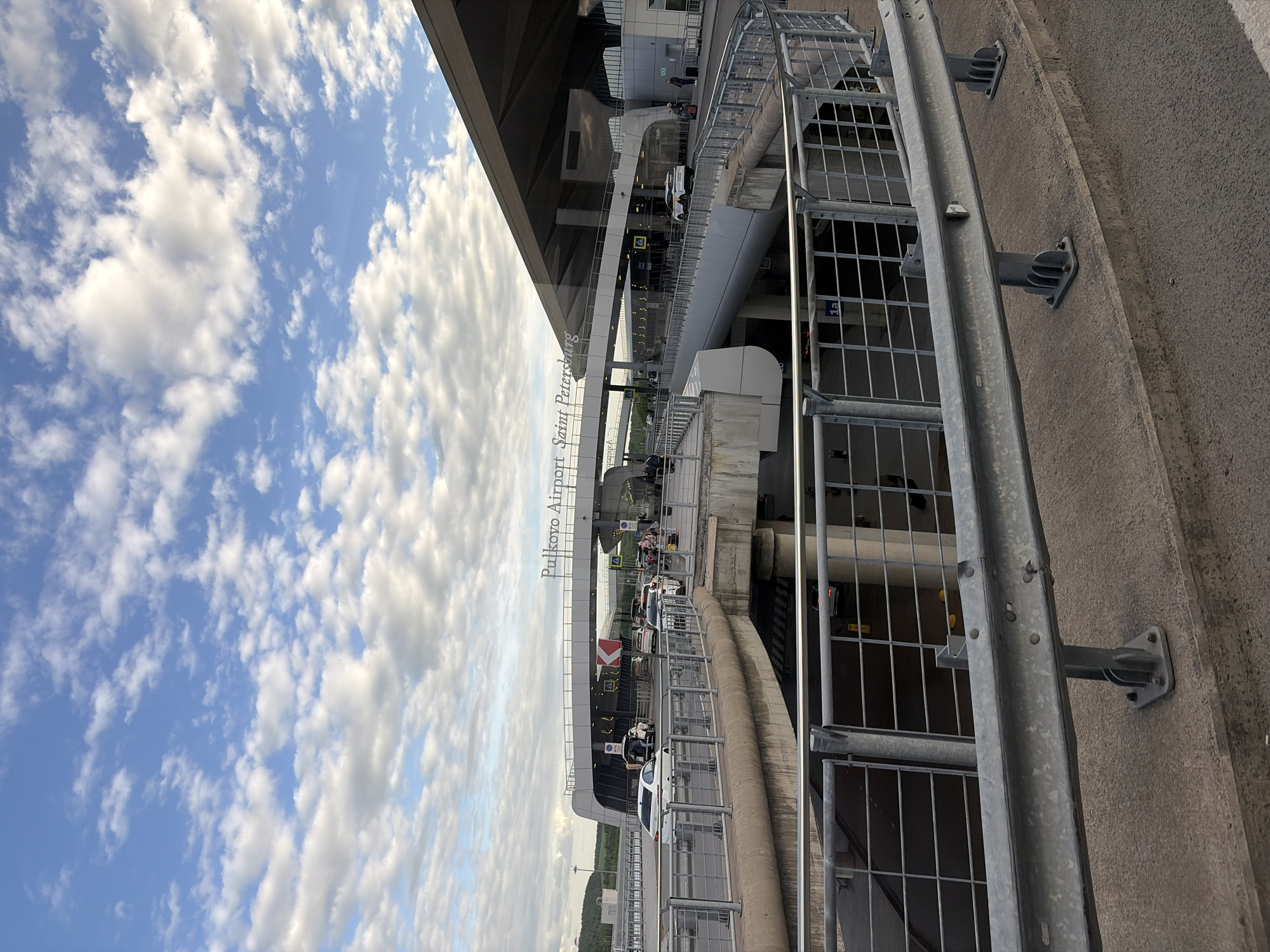 The image depicts Pulkovo Airport in Saint Petersburg, Russia. The view is from an elevated position, likely a parking structure or overpass, looking towards the airport terminal. A metal guardrail is prominent in the foreground on the right. The airport building is visible with glass panels and overhang. Cars and people are observable near the entrance. The sky is filled with cumulus clouds.