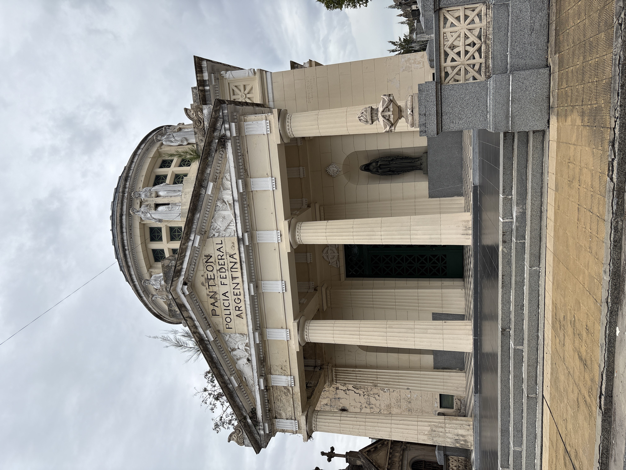 The image depicts the Pantheon of the Argentine Federal Police, located within the Recoleta Cemetery in Buenos Aires, Argentina. The prominent structure is a large, classical-style mausoleum, characterized by a light beige facade that shows signs of weathering and discoloration.
The main entrance features a portico supported by four tall, fluted columns, leading to recessed dark green double doors with a grid pattern. Above the columns, a triangular pediment contains a bas-relief sculpture depicting figures and heraldic elements. The facade also displays the inscription "PANTEON POLICIA FEDERAL ARGENTINA" in relief lettering. To the right of the entrance, a dark, robed female statue stands within a shallow niche.
The mausoleum is crowned by a multi-tiered dome, adorned with several lighter-colored allegorical statues positioned around its base. Small plants are observed growing from crevices and ledges on the structure, particularly near the pediment and dome.
In the foreground, a series of dark grey stone steps leads up to a reflective black tiled platform directly in front of the mausoleum's entrance. The immediate ground surface is paved with tan-colored, patterned tiles. To the right of the steps, a low grey stone railing features a decorative geometric lattice infill panel and is topped by a decorative urn-like finial.
The environment is an overcast sky, with no direct sunlight. In the background, particularly to the left and right, numerous other elaborate mausoleums, crosses, and tombstones are visible, confirming the cemetery setting. No individuals are present in the scene.