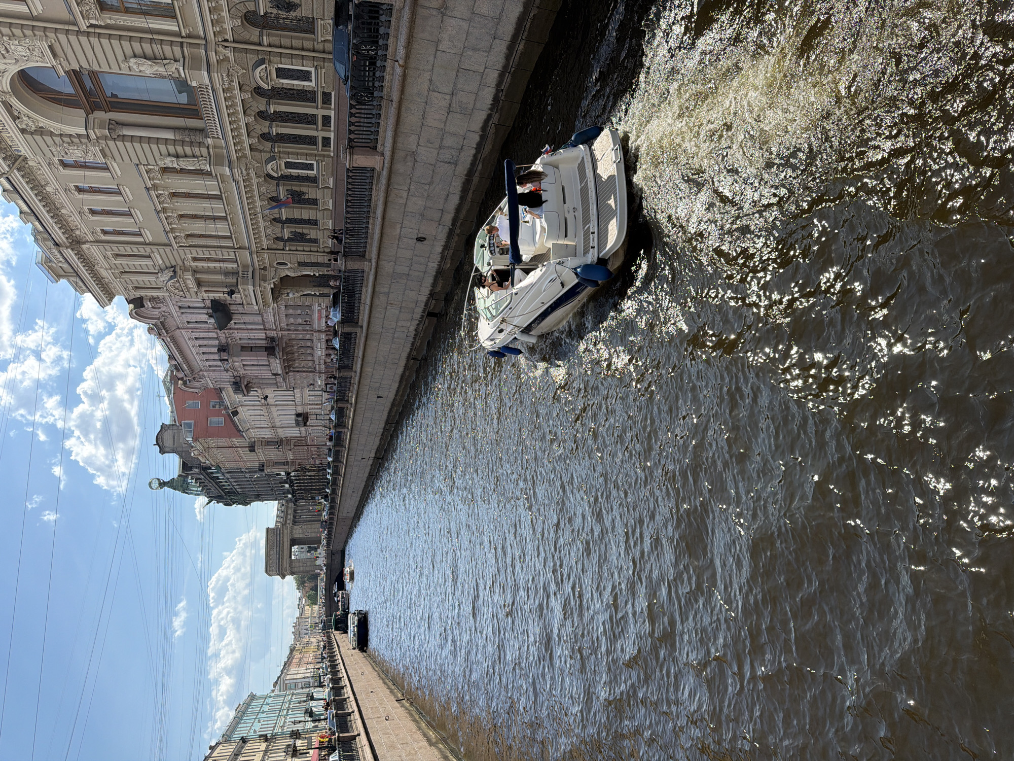 The image, taken from an elevated perspective, captures a scenic canal in Saint Petersburg, Russia, on a bright, sunny day. The canal, filled with dark, rippling water reflecting sunlight, stretches into the distance, flanked by grand, ornate buildings.
In the mid-ground, a white motorboat with blue fenders navigates the canal, leaving a foamy wake behind it. Several people are visible on board the boat, appearing to enjoy the ride; at least three individuals can be clearly seen, including one person standing near the bow and others seated. A small Russian flag is mounted on the stern of the boat.
The left bank of the canal is lined with a majestic, richly decorated building featuring intricate architectural details, such as pilasters, sculptures, and ironwork balconies. Above a large arched window on this building, faint Cyrillic text can be partially deciphered, appearing to read "МАГАЗИН" (Shop) and possibly "ДЕНЬ И НОЧЬ" (Day and Night) below it. Further down the embankment, parked cars are visible.
On the right bank, other buildings of varying architectural styles and colors can be seen, leading to a distant archway or bridge. The sky above is a clear blue with scattered white, fluffy clouds. Numerous power lines or cables crisscross the upper portion of the sky, adding a modern element to the historic cityscape. The overall atmosphere is peaceful and leisurely, characteristic of a pleasant day spent sightseeing by water.