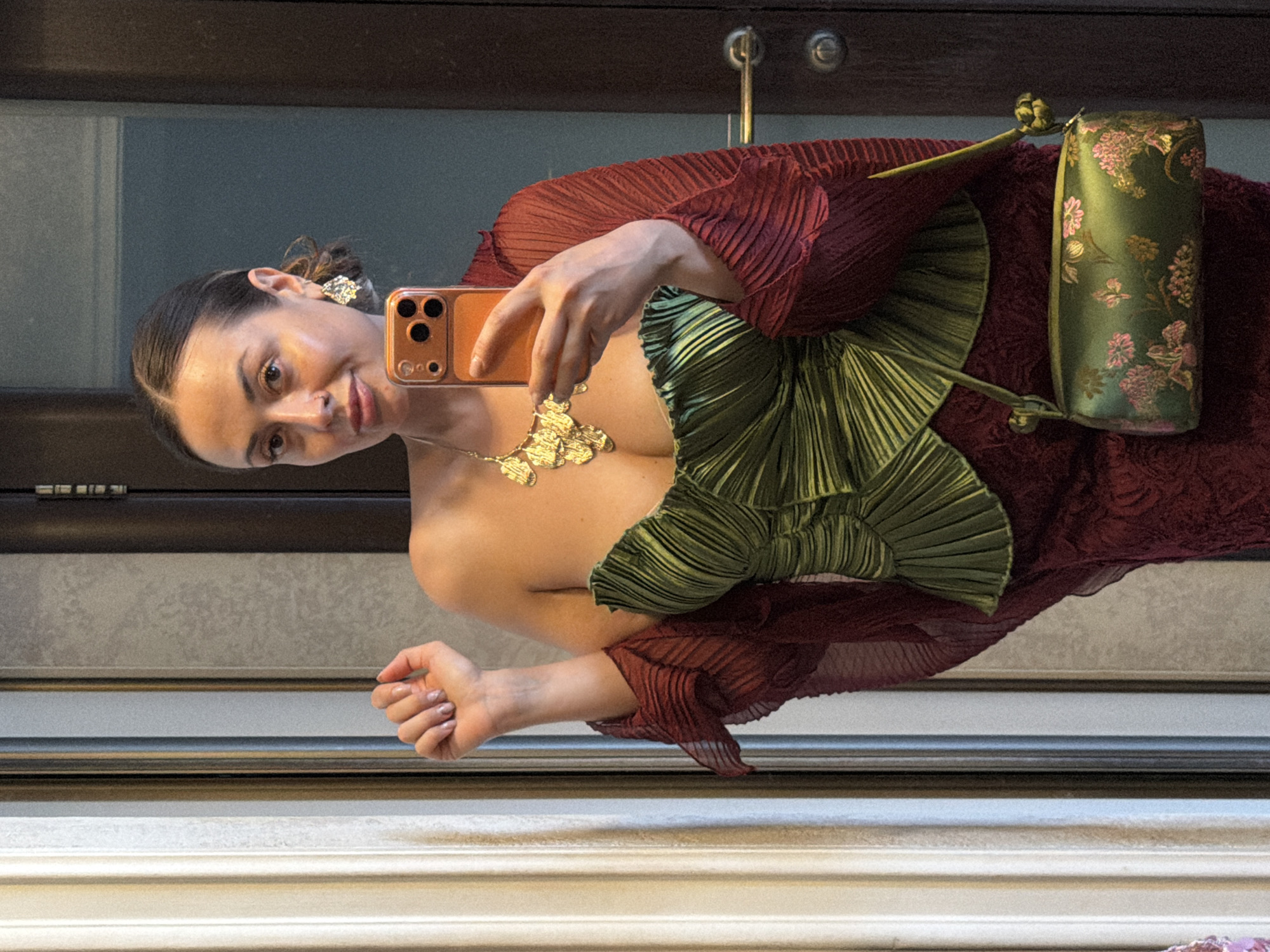 A woman stands in a dimly lit room, taking a mirror selfie. She is wearing a green, pleated, strapless dress and a dark red, sheer shawl draped over her shoulders. She accessorizes with a large gold necklace and dangling earrings. A green, embroidered clutch purse hangs from her left shoulder. She holds an orange smartphone in her right hand, capturing her reflection. The background consists of a reflective surface, likely a mirror, which shows a doorway and a ribbed curtain or panel. Based on the available information and common architectural features, this scene could be located in an apartment or a boutique in Krasnoyarsk, Russia.