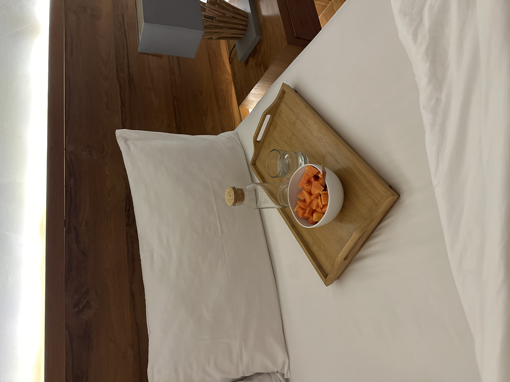 The image shows a wooden tray with a bowl of cut fruit, a glass, and a water bottle with a cork stopper, resting on a white bed. A white pillow is positioned next to the tray. The background reveals a wooden headboard and bedside table. The location is likely a hotel or residence in Sukawati, Indonesia, given the style and décor. There are no people present in the scene.
