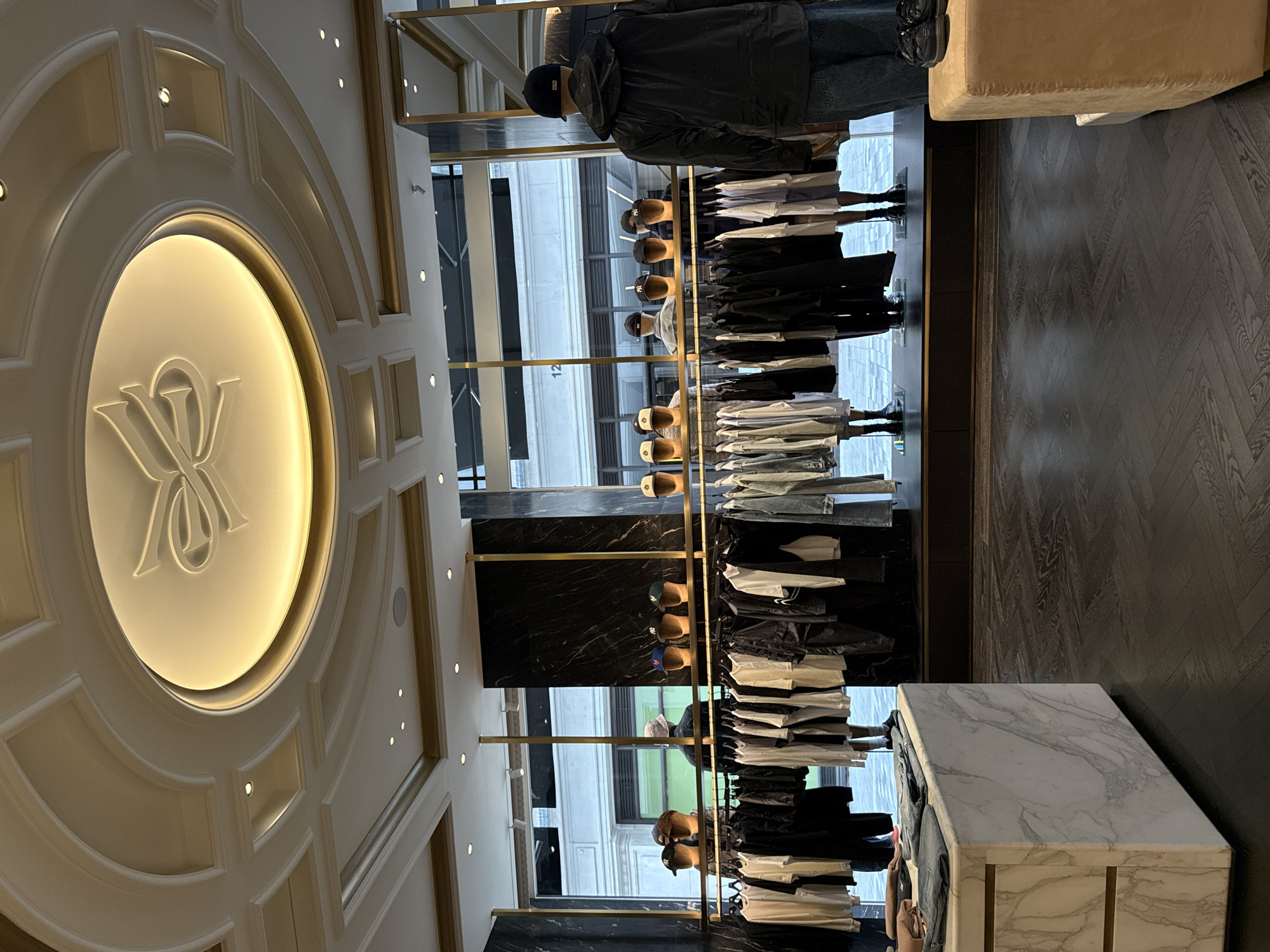 The image displays the interior of an upscale retail clothing store in London, United Kingdom. The floor is dark wood laid in a herringbone pattern. The ceiling is white and ornate, featuring recessed lighting and a prominent central circular fixture with an illuminated perimeter, inside which is a raised, stylized emblem resembling a looped letter 'K' or an intertwined ribbon.
Large floor-to-ceiling windows form the background, offering a view of an urban street and a light-colored building with classical architectural elements across the way; the number '12' is visible on one of the building's windows. These windows are flanked by dark, veined marble panels and accented with gold-colored metal fixtures.
In the mid-ground, multiple clothing racks with gold-toned rails are arranged, displaying an assortment of garments primarily consisting of t-shirts, hooded tops, and jeans in a color palette of white, black, grey, and denim blue. Several headless mannequins stand behind these racks, some wearing baseball caps, including caps with the New York Yankees logo. On the right side of the image, a full-figure mannequin is visible from the back, dressed in a black hooded jacket and blue jeans. Adjacent to this mannequin, a pair of black shoes rests on a beige upholstered cube, which appears to be a display or seating element. In the left foreground, a large rectangular display table made of white marble with distinct grey veining holds neatly folded denim jeans and other apparel. No live individuals or active interactions are visible within the scene.