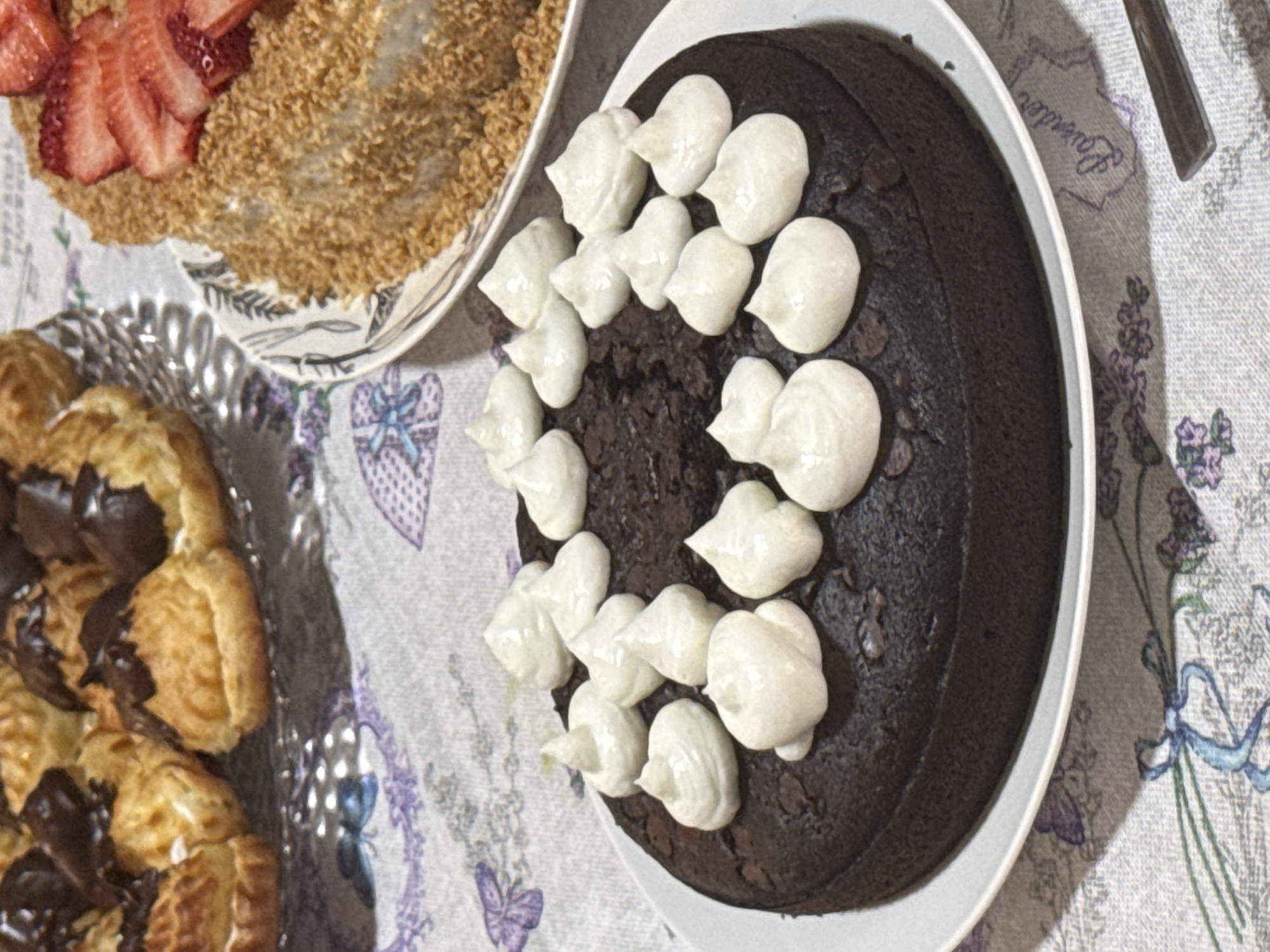 A table setting with several desserts. On the right is a round chocolate cake with white frosting arranged in a circle, sitting on a white plate. To the left of it is another cake, possibly layered, topped with sliced strawberries and a light-brown grainy topping, on a white plate with a decorative pattern. In the lower left corner is a silver platter filled with cream puffs drizzled with chocolate. The tablecloth has a light-colored background with a pattern of lavender and butterflies. A knife is near the chocolate cake. The location is not discernible from the image.