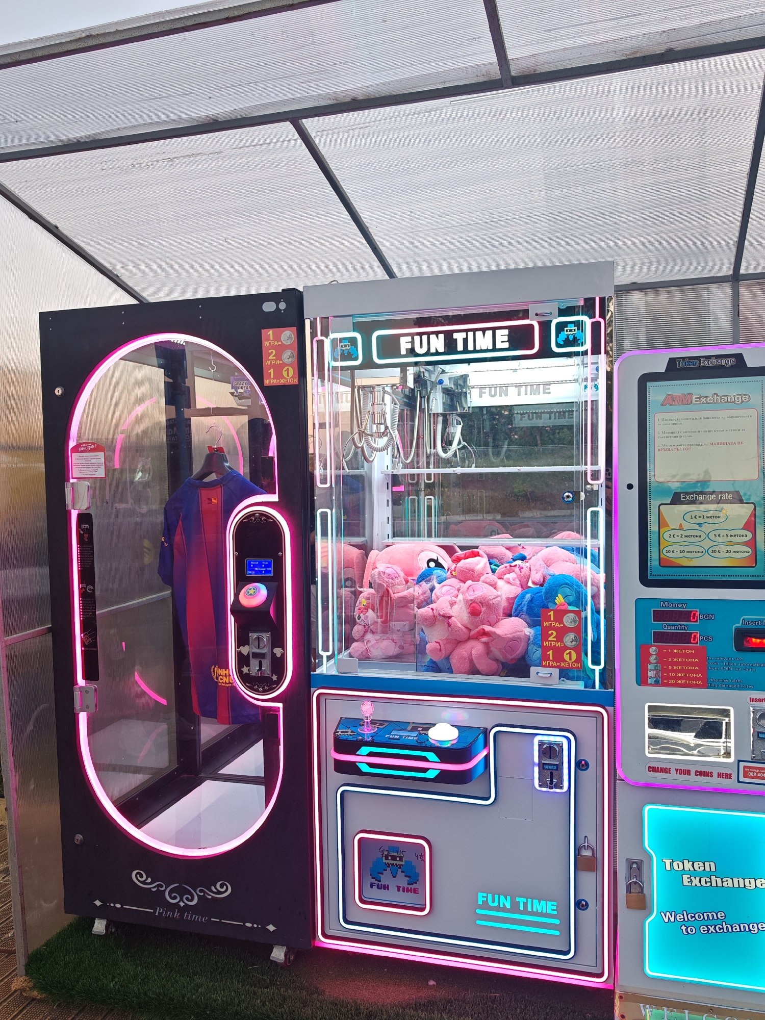 The image presents a set of amusement claw machines under what appears to be a translucent awning or roof structure, likely in an outdoor setting. These machines are indicative of an arcade or entertainment area. One machine, labeled "Pink Time", is black with pink neon accents, featuring a blue and red Barcelona jersey inside. The central machine is branded "Fun Time", with a brightly lit sign and a claw crane over an assortment of stuffed animals in pink and blue. The machine on the right is a "Token Exchange" unit, with a screen providing exchange rates and instructions in Bulgarian. The setting is possibly in Nesebar, Bulgaria.