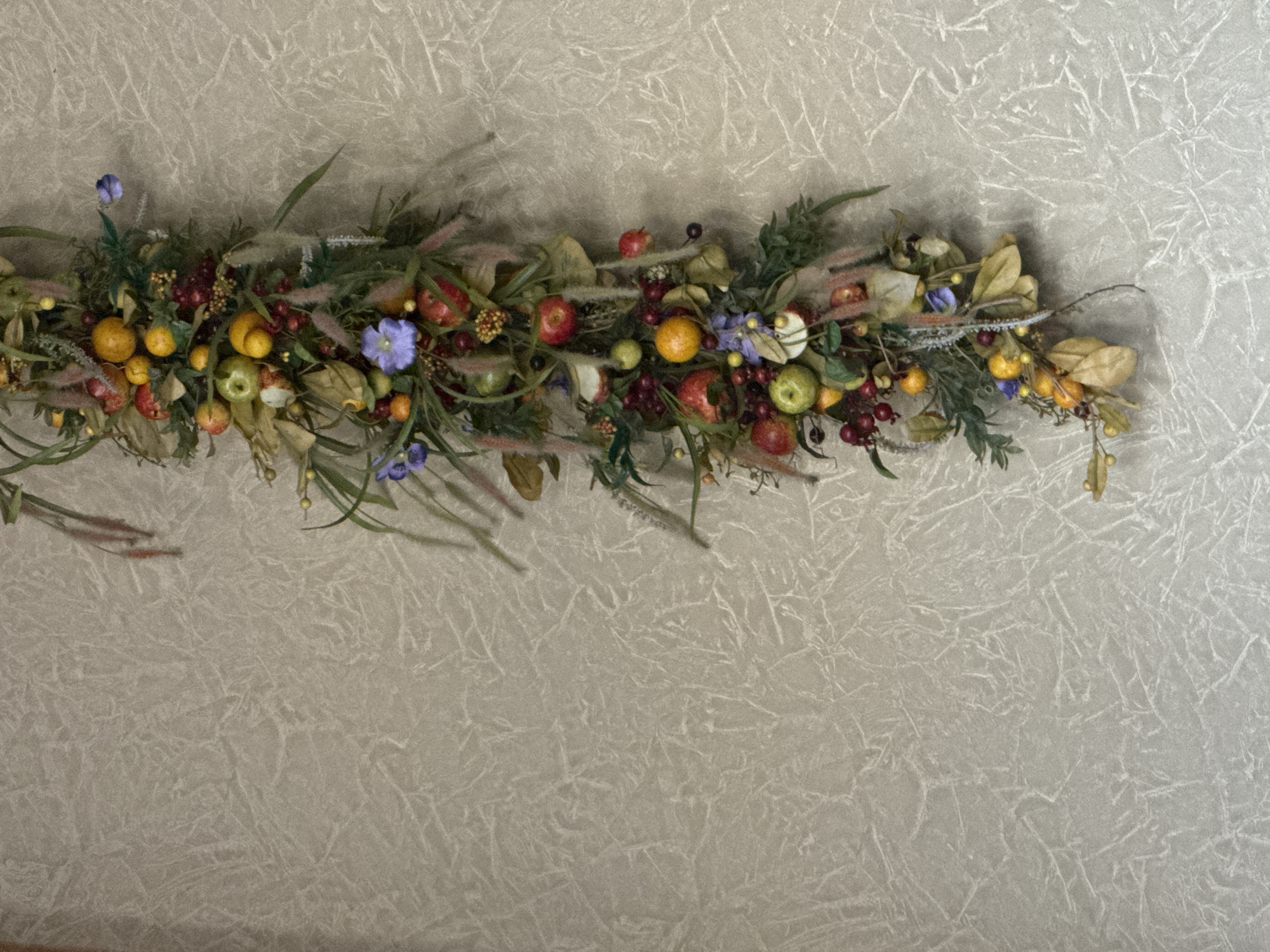 A vertical, artificial fruit and foliage garland is attached to a textured, light-colored wall. The garland is composed of various faux fruits, including apples, oranges, and berries, interspersed with green leaves, dried foliage, and small purple flowers. The garland hangs from the top right of the frame and extends downwards along the right edge of the image. The background wall has a subtle, irregular pattern resembling cracked plaster or a distressed finish. The lighting appears to be artificial and diffused, casting no harsh shadows. The scene is static and appears to be an indoor decoration. There are no people or animals present. The location in Zaokskiy, Russia, is not visually represented in the image.
