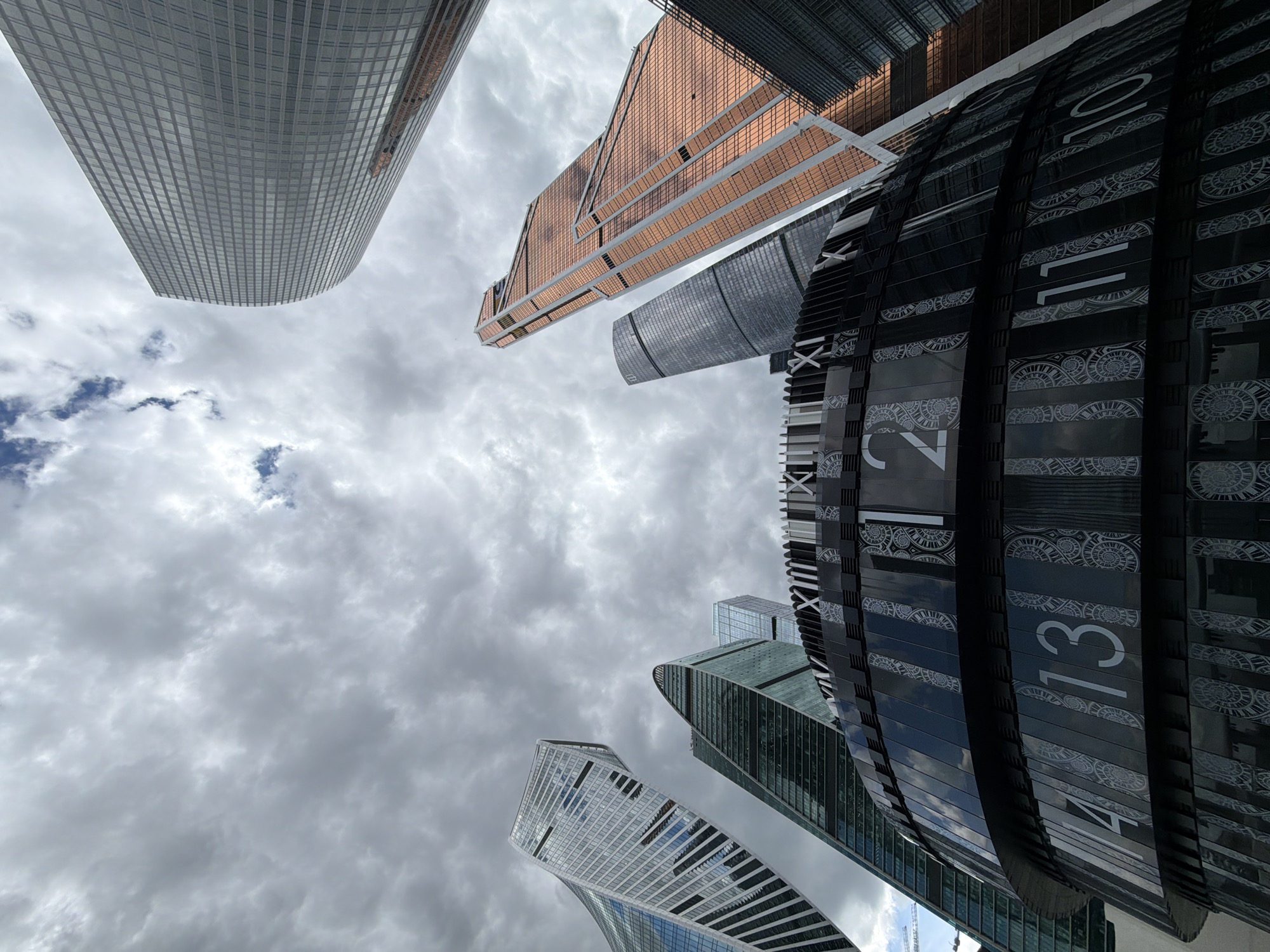 The image captures a low-angle view of a cluster of modern skyscrapers set against a cloudy sky. The buildings vary in design and materials, including glass, metal, and stone. Prominent in the foreground is a dark, cylindrical structure featuring numbers and decorative patterns. The sky is predominantly overcast with varying shades of gray clouds, with small patches of blue visible. The location appears to be a modern urban area with high-rise architecture.