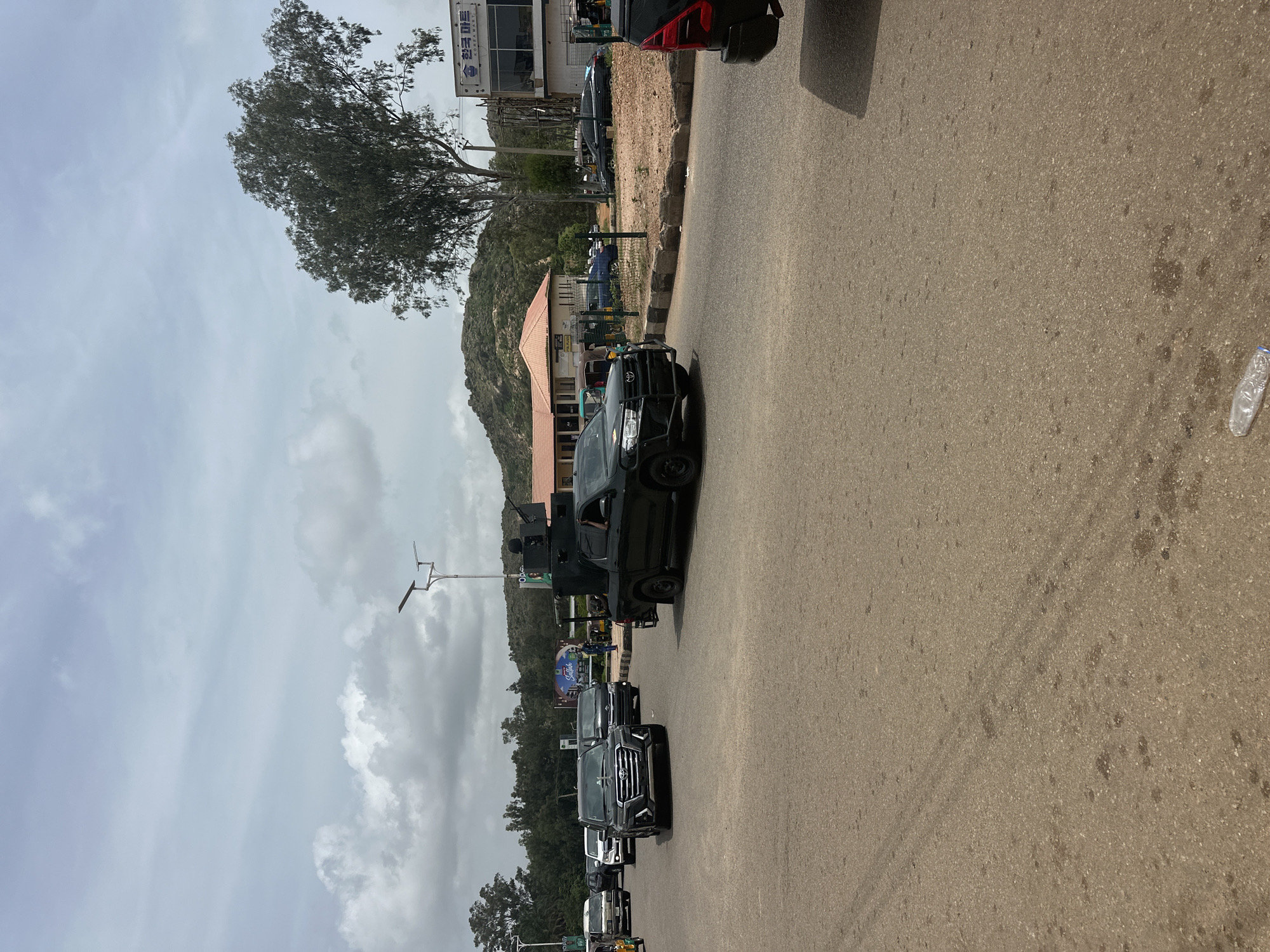 A motorcade, likely a security detail, moves along a paved road in Gura Top, Nigeria, under an overcast sky. The lead vehicle is a black Toyota Land Cruiser with a reinforced front bumper and tinted windows, followed by several other dark-colored SUVs. To the right, a black pickup truck with a mounted machine gun and at least two visible occupants, one possibly armed, is prominent. In the background, buildings with red tile roofs and a taller structure with Korean signage ("한국 마트," Korean Mart) are visible. The environment includes sparse vegetation, rocky hills, and a street light. The road surface shows signs of wear and occasional debris, including a discarded plastic bottle.