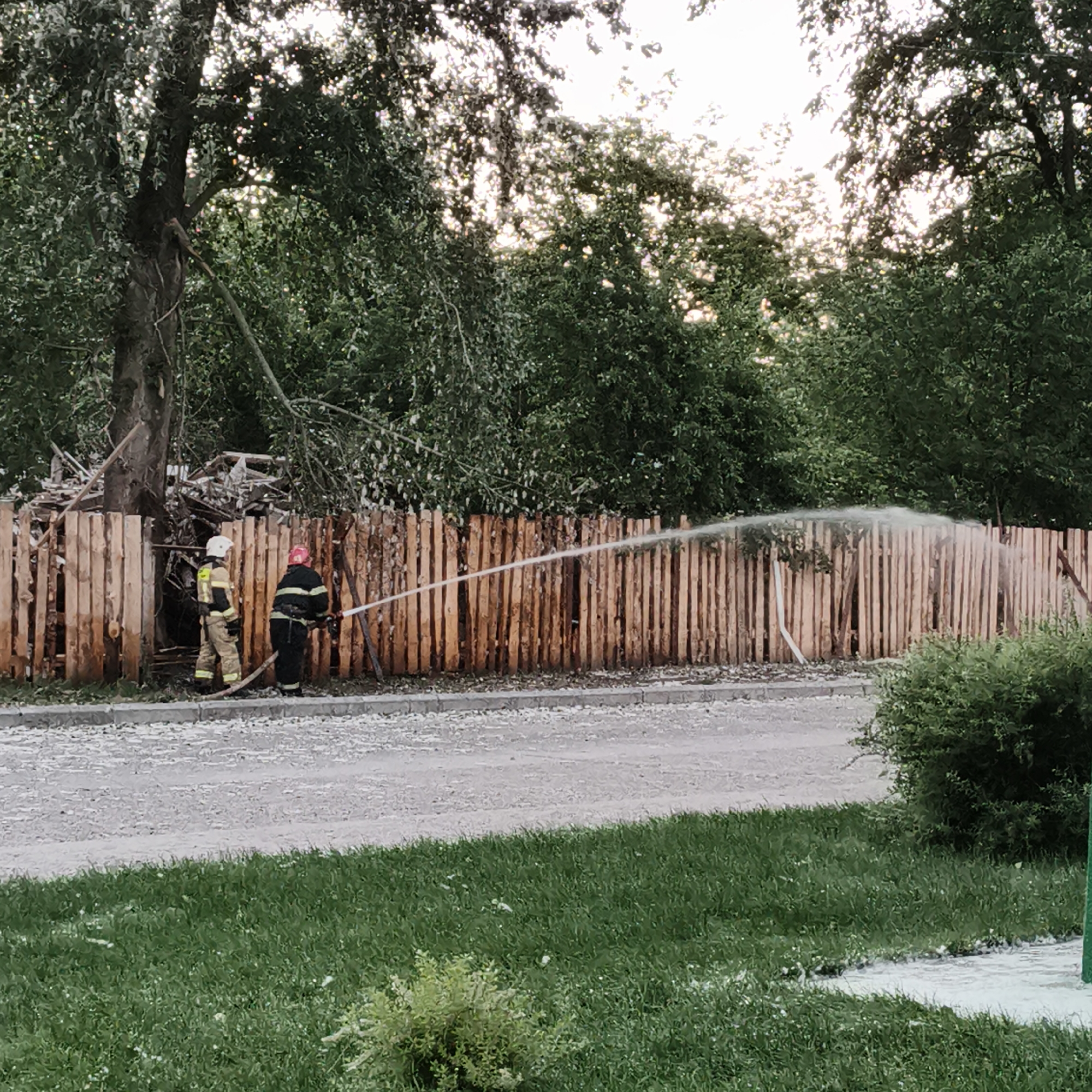 In Chaikovsky, Russia, two firefighters are seen in action near a wooden fence. One firefighter, wearing a white helmet and a tan uniform, stands to the left. The other, with a red helmet and a black and yellow striped uniform, is to the right, operating a hose that sprays water towards the fence. Behind the fence, debris and a fallen tree are visible, suggesting a recent incident. The scene is set outdoors with lush green trees and grass surrounding the area, under an overcast sky. The ground in front of the fence is covered with gravel and small white objects, possibly remnants from the incident.