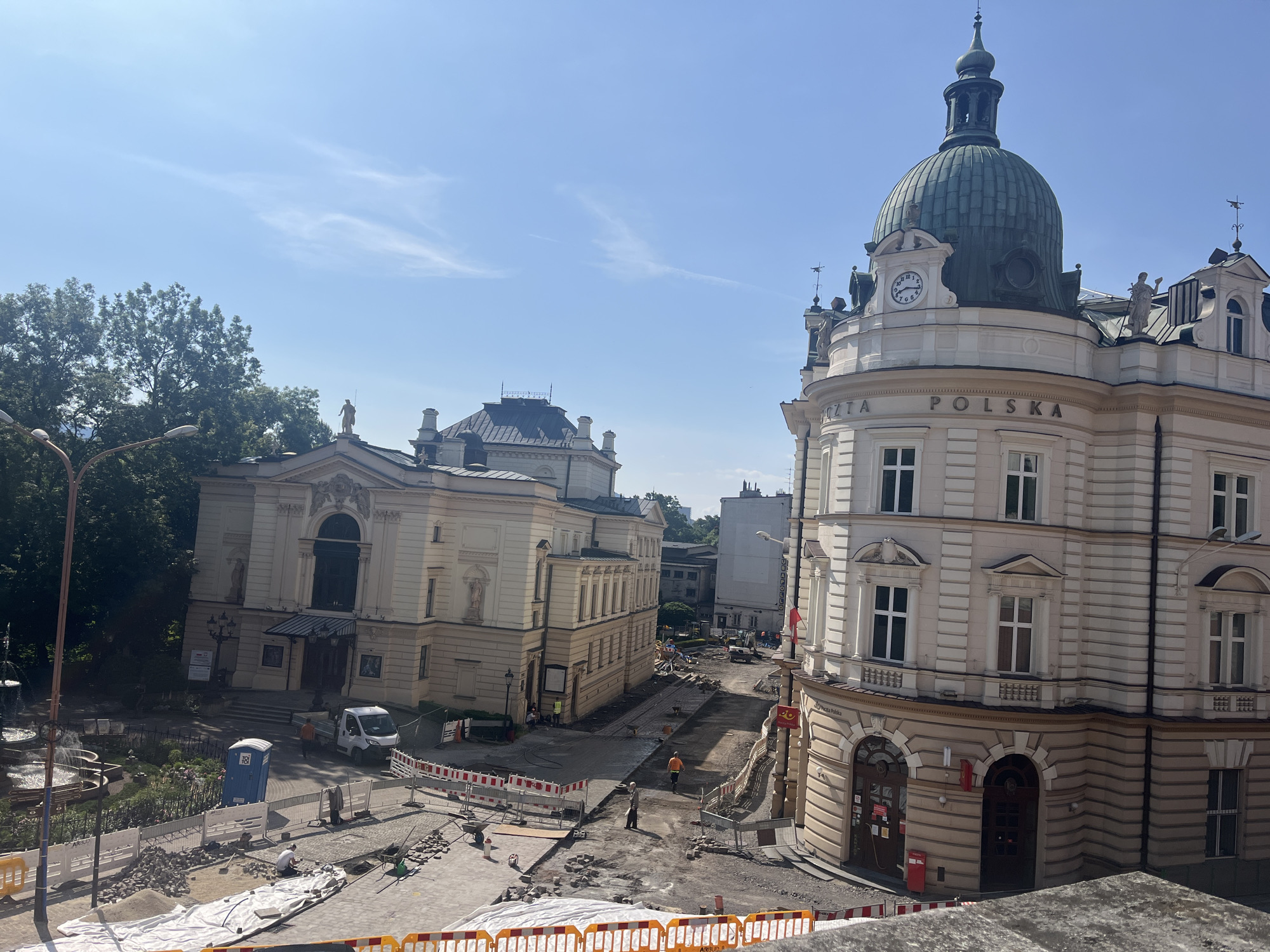 The image shows an outdoor scene of a city street under construction. In the foreground, there are construction barriers and piles of materials. In the middle ground, several buildings are visible, including a large, ornate building with a green dome and a clock, and a smaller, tan-colored building that appears to be a theater or opera house. Workers in construction gear are present, some engaged in labor, while others appear to be supervising. A white van is parked near one of the buildings. The street itself is partially paved, with areas of exposed dirt and cobblestones. The background features trees and more buildings, indicating an urban setting. The sky is blue with a few wispy clouds.
