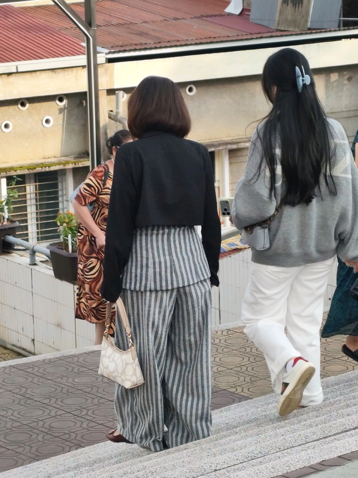 In Da Lat, Vietnam, three individuals are captured on an outdoor staircase. The first, a woman facing away from the camera, wears a black cropped jacket over striped gray and white wide-leg pants, carrying a cream-colored handbag with a patterned strap. Her dark hair is cut to shoulder length. To her right, another woman also faces away, her long dark hair secured with a light blue clip. She is dressed in a loose-fitting gray sweater with a subtle pattern and white pants, carrying a small light blue crossbody bag and holding a smartphone. Behind them and slightly to the left, a third woman, partially obscured, wears a brown and black animal-print dress. The staircase features a patterned gray tile tread and a rough, speckled riser. The background shows a building with a red corrugated metal roof and light yellow walls, featuring rows of circular vents. A potted plant sits on a ledge to the left of the staircase. The lighting suggests it is daytime, possibly late afternoon given the shadows.