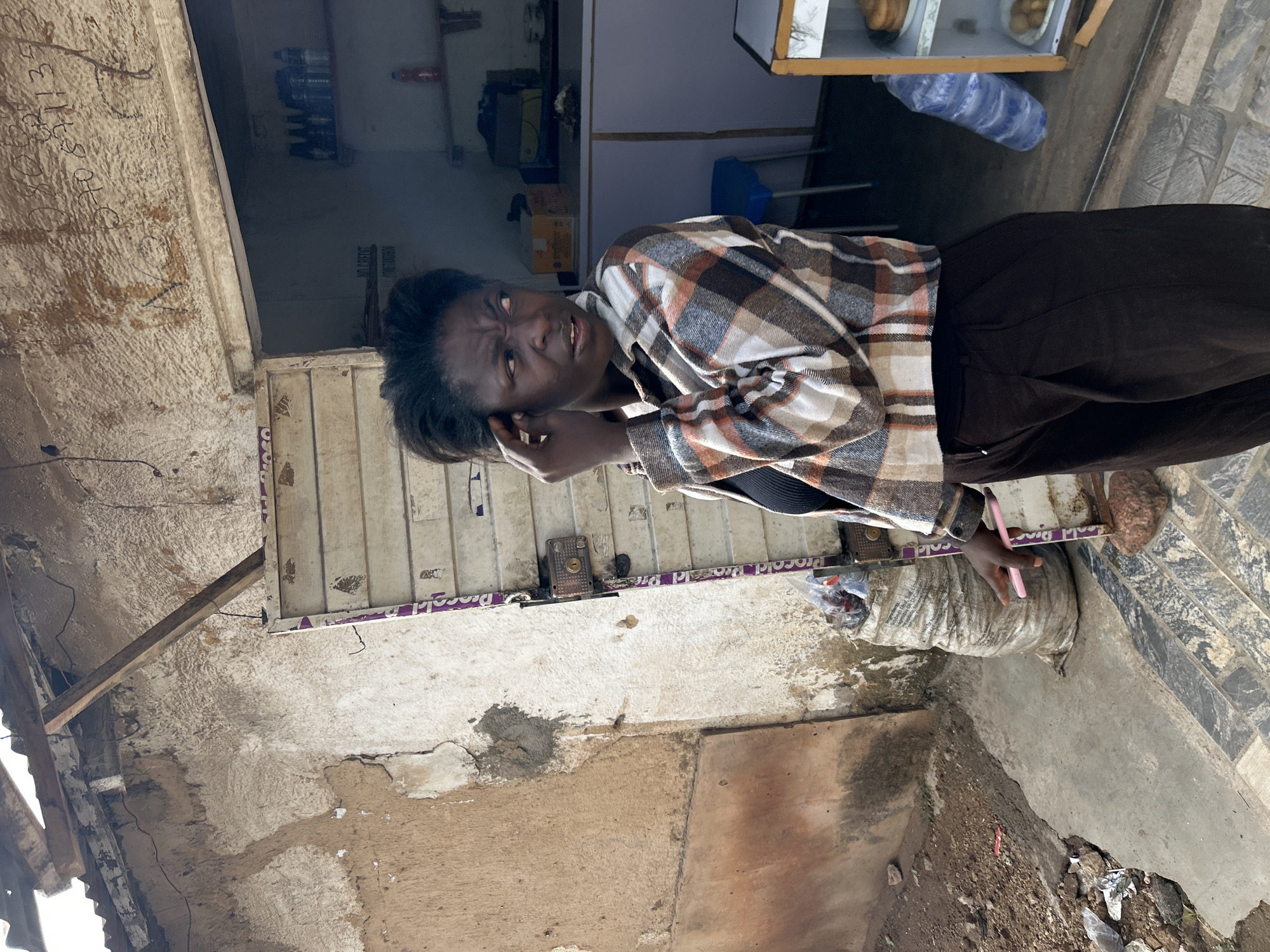 A woman is outdoors in Jos, Nigeria. She's wearing a plaid shirt and dark pants, holding a pink smartphone to her ear. She appears to be in front of a small store or building with a cluttered interior visible through an open doorway. The building's exterior is worn with visible damage. A large bag sits on the ground near the woman. A food display is visible over her head. The ground is concrete or stone, with trash and debris scattered nearby.