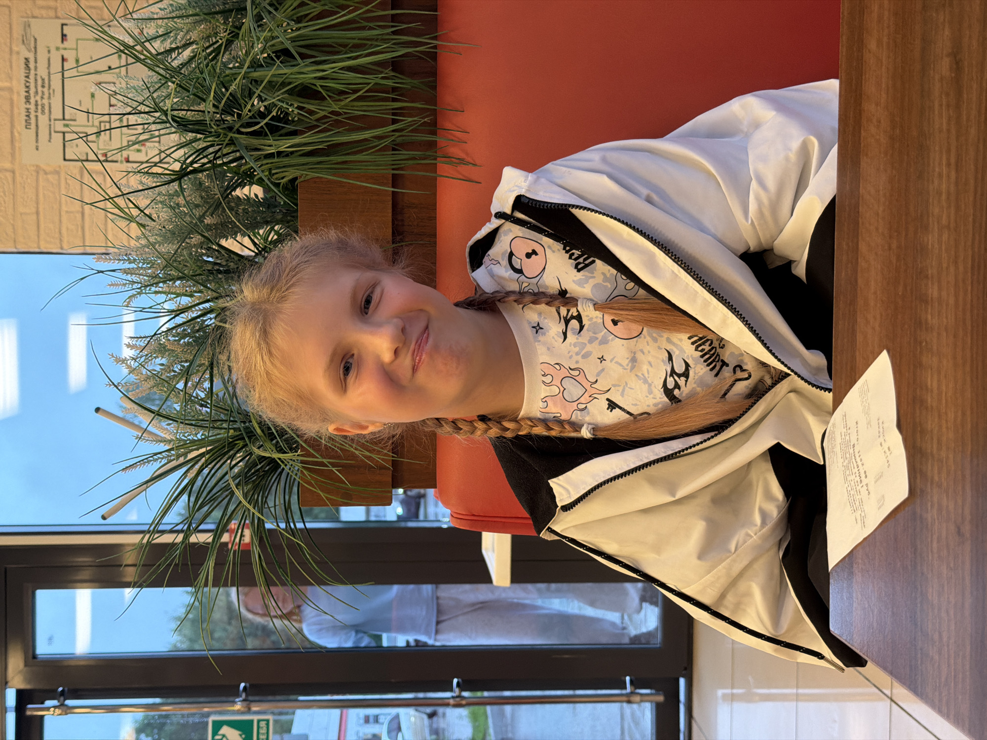 A young girl with blonde hair in pigtails is shown from the chest up, sitting at a wooden table. She is wearing a white jacket over a t-shirt with a cartoonish print featuring pink hearts and musical notes. Her left hand is visible, resting on the table. To her left, a large, leafy green plant is visible. Behind her, a red booth seat is present. In the background, through a glass window, a reflection of people in light-colored clothing can be seen, along with a glimpse of an outdoor area with a blue sky, suggesting daylight. A sign with Cyrillic text and a diagram is affixed to a light-colored wall in the upper left quadrant of the image. A white receipt is placed on the table to the right of the girl, partially obscured by her jacket. The scene appears to be indoors, possibly a restaurant or cafe, in Krilovo, Russia. The girl has a cheerful expression.