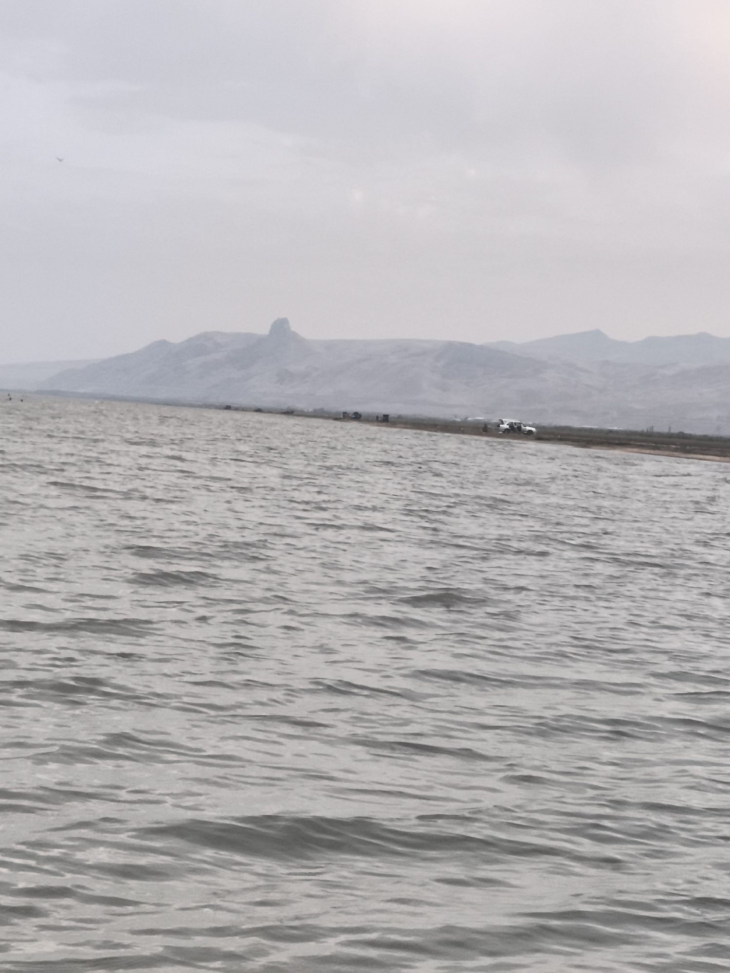 This is a horizontal, medium-shot photograph of a body of water with a distant, hazy mountain range. The water in the foreground is rippled and appears grey under an overcast sky. Along the far shoreline, a thin strip of land is visible, with several vehicles, including a white pickup truck with its doors open, parked along it. Further in the distance, the hazy mountain range has a distinct, jagged peak towards the left. The sky is a uniform, light grey, with a faint hint of sun towards the upper right. A single bird is visible in the upper left corner of the frame. The scene appears to be a natural landscape, potentially near Balaca Həmyə, Azerbaijan, given the mountainous terrain.