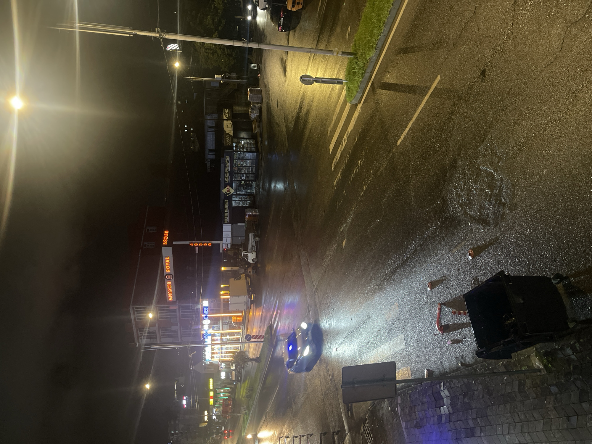 The image depicts a wet street at night in Batumi, Georgia. A blue car with flashing lights is driving on the wet asphalt road, with reflections of streetlights and building lights visible. Several buildings with illuminated signs are located on the right side of the street. One building has a large sign that reads "ROYAL" in red letters, with Georgian text above it. Another building has signs for "BUILDING MATERIALS". There are streetlights and power poles along the side of the road. A pedestrian crossing is marked on the road. In the foreground, a black trash bin is visible on the sidewalk, with traffic cones and other debris scattered nearby. The overall scene is dark, with strong artificial lighting creating reflections and highlights on the wet surfaces.