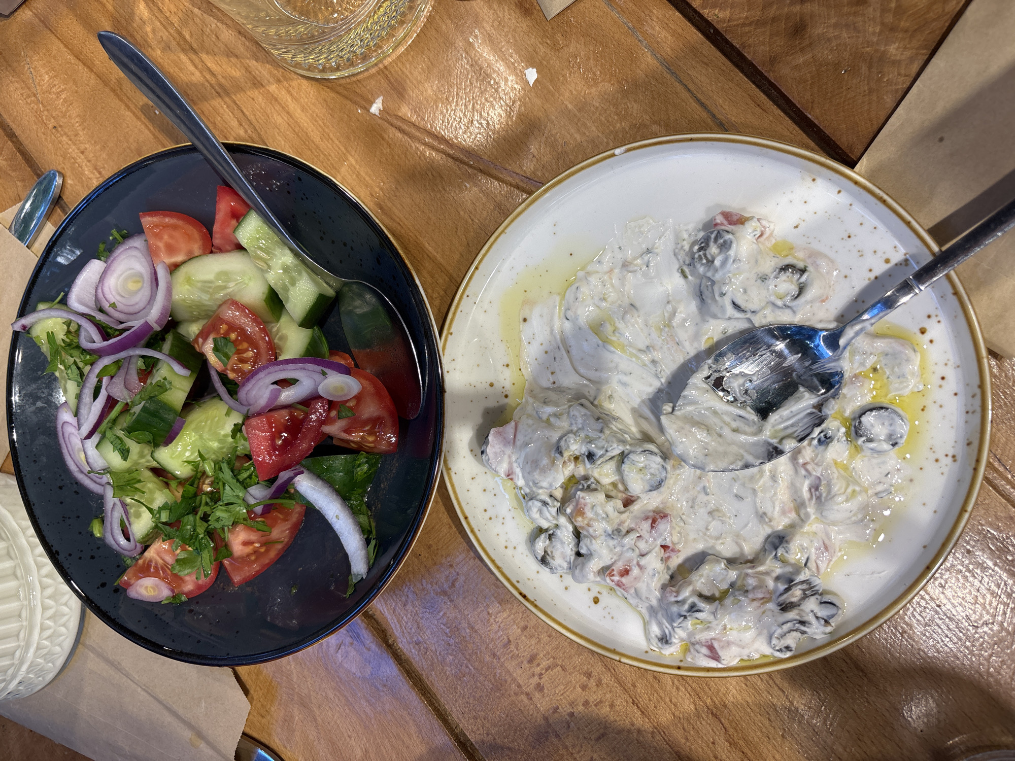 A top-down shot shows two plates on a wooden table, likely in a restaurant in Yerevan, Armenia.
The left plate, a dark grey with a blue rim, holds a mixed salad. The salad contains sliced cucumbers, red tomatoes, red onion rings, and chopped green herbs, possibly parsley. A serving spoon rests within the salad.
The right plate, white with a speckled brown rim, contains a partly consumed dip or salad, possibly a yogurt-based sauce with ingredients mixed in. A spoon is visible in the plate. A light-colored oil appears to be drizzled over the dip.
The wooden table has a striped pattern. In the background, a partially visible glass with a decorative texture and a brown paper surface are present.