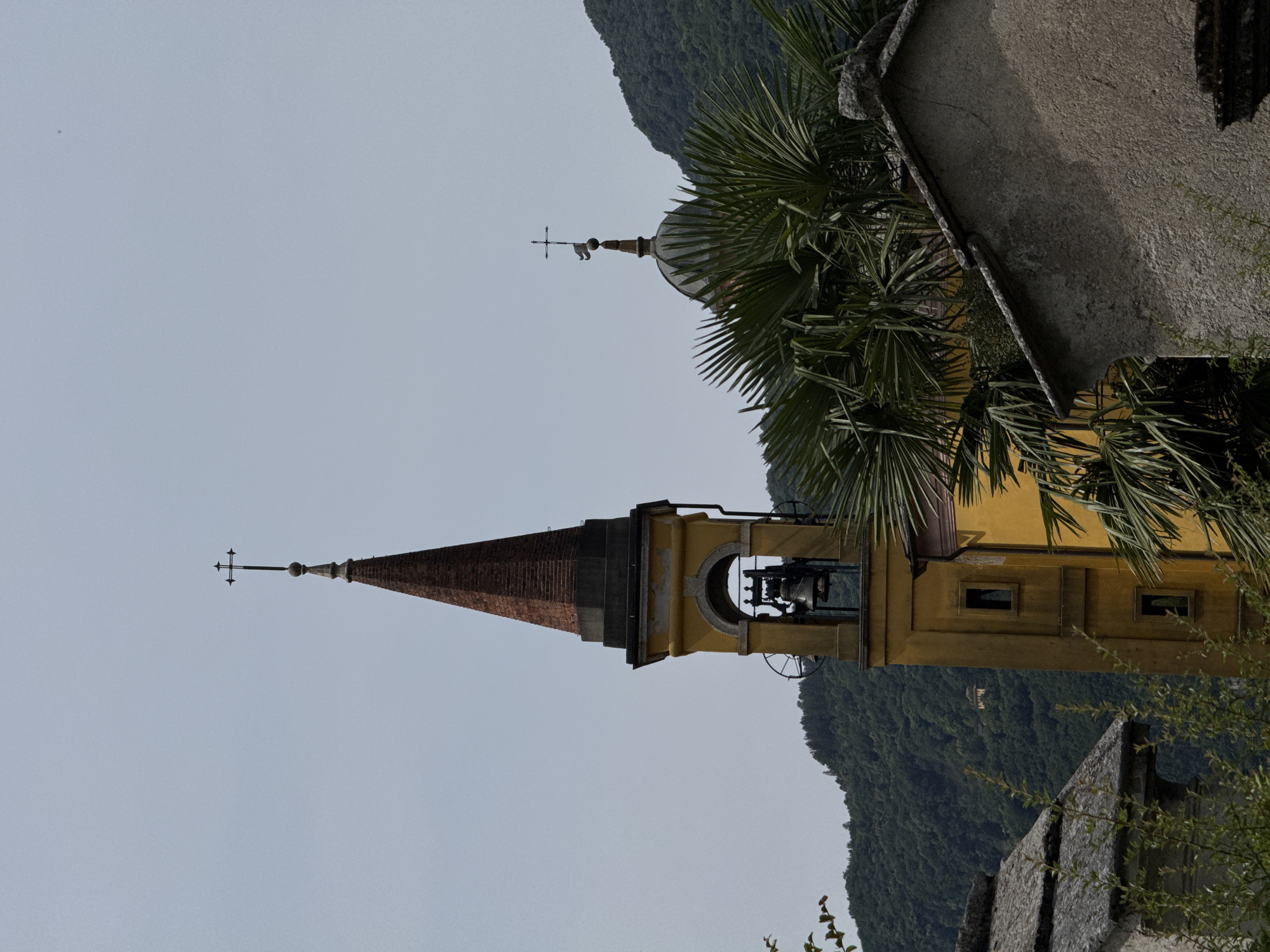 A yellow church tower with a red-tiled, conical roof stands prominently against a pale sky. A cross tops the spire. The tower has arched openings revealing a bell and associated machinery. To the right, a domed structure, also topped with a cross and a weather vane, is partially visible, obscured by lush green palm fronds. In the background, a densely forested mountain slopes upward. In the foreground, stone buildings and foliage partially frame the scene.