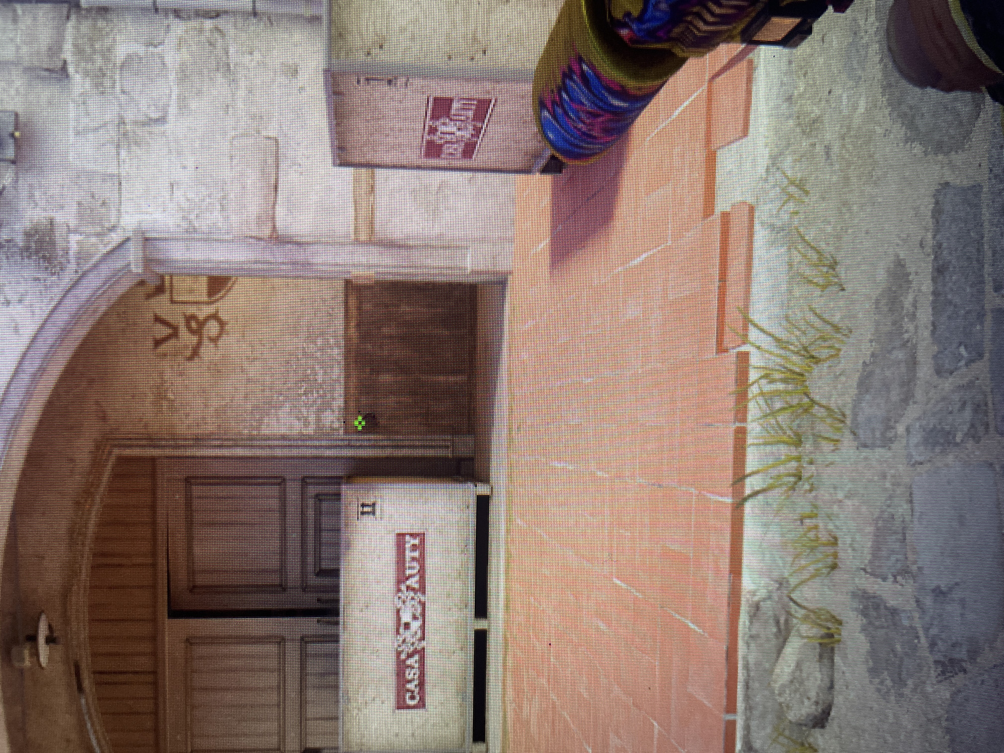 The image depicts a first-person perspective of a video game environment, likely from a first-person shooter. The scene appears to be set in an outdoor courtyard with terracotta tiled flooring and stone walls. In the background, there are large wooden doors and architectural elements suggesting an old building or structure. There are also crates with logos that are partially visible. The player's hand holding a weapon with a colorful patterned grip is visible in the foreground. The image does not provide any information or visual cues related to the city of Kołobrzeg, Poland.