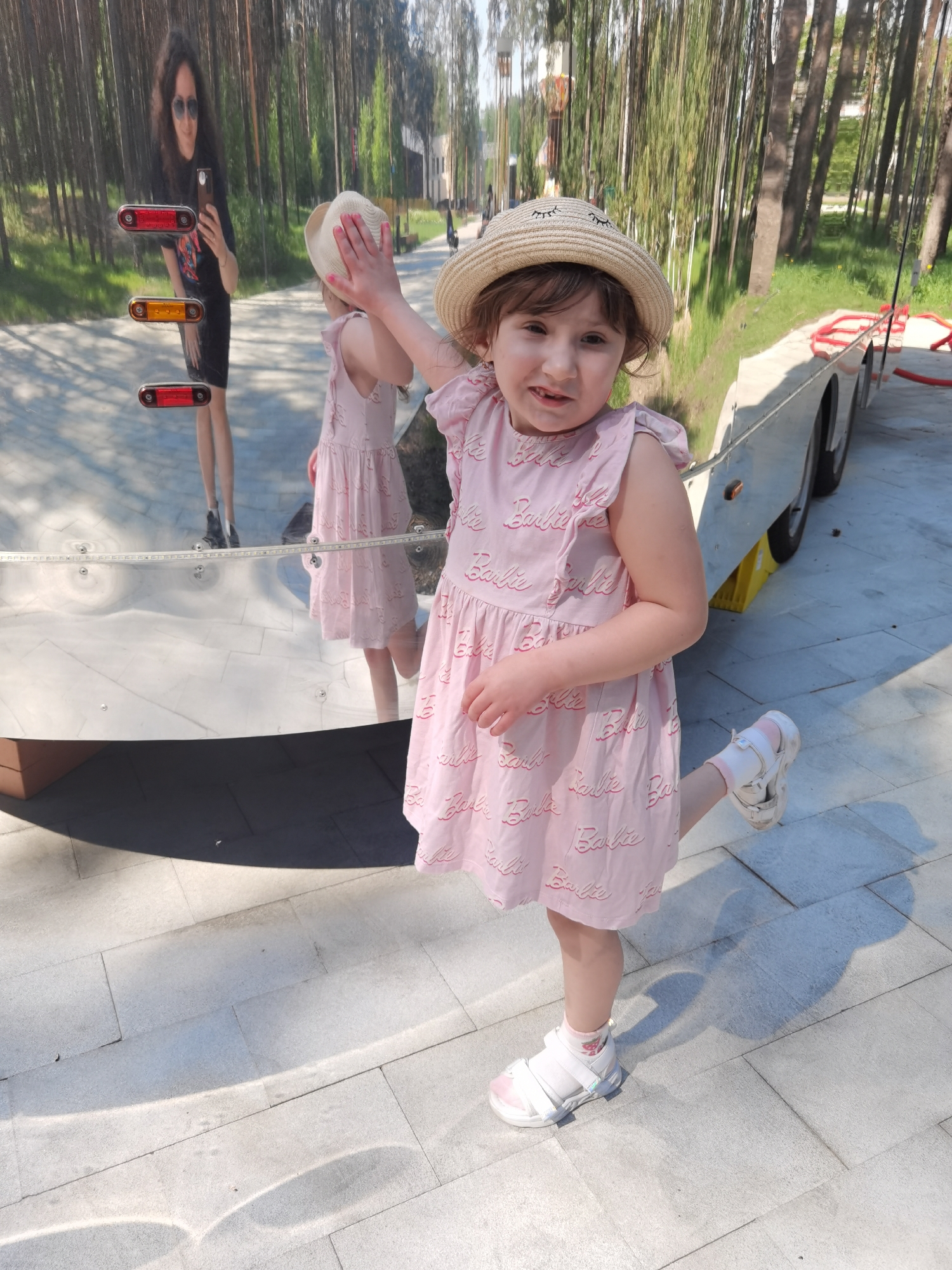 A young girl in a pink Barbie dress and a straw hat is standing on a paved area, possibly a sidewalk, with one leg raised. She is posing in front of a mirrored surface that reflects the surrounding environment, including trees and a woman taking a selfie. The location is identified as Perm, Russia, likely in a park or public area with trees and art installations. There is also a utility trailer next to the girl.