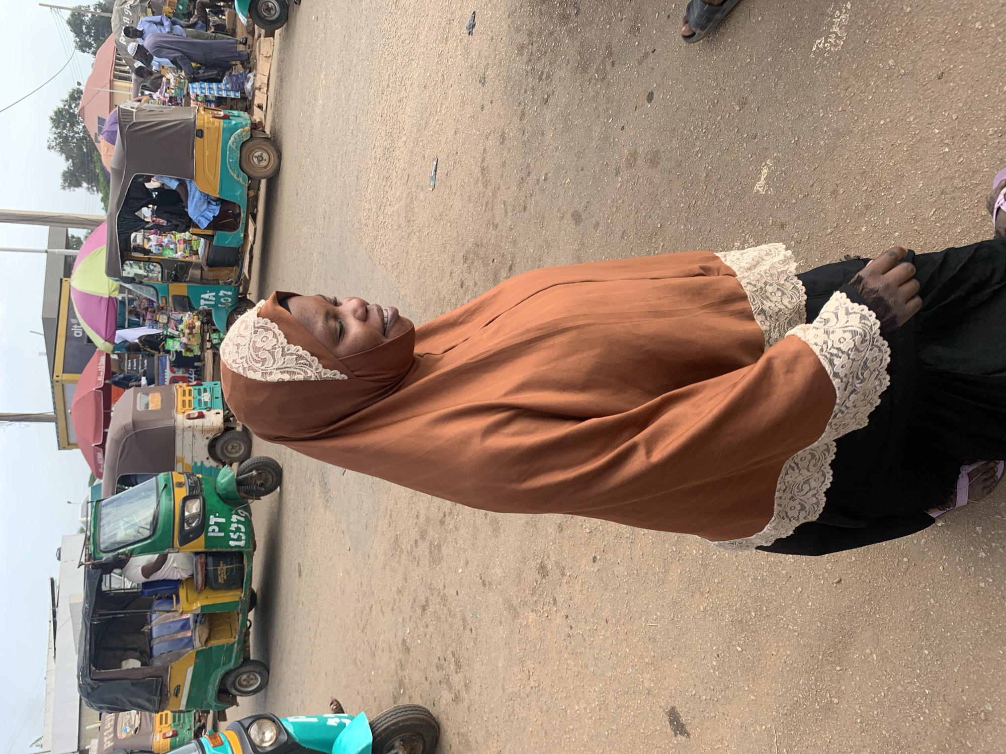 In Gura Top, Nigeria, a woman wearing a brown hijab with white lace trim and a black skirt kneels on a dirt road. She is looking to her right with a slight smile. In the background, several colorful auto-rickshaws, also known as "Keke Napep," are parked or driving on the road. One auto-rickshaw in the foreground is green and yellow with "PT 1537" written on its side. Another auto-rickshaw to the right is yellow and teal with "PTA 107" visible. People are standing around the auto-rickshaws and in the background, some appearing to be vendors. The environment appears to be a market or a busy street with stalls set up. The sky is overcast.