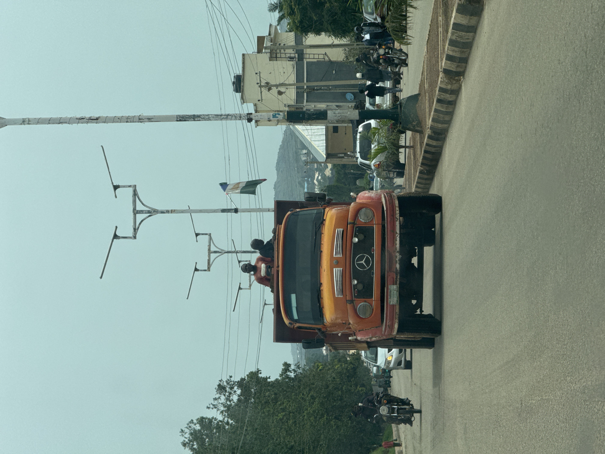 The image shows a street scene in Gura Top, Nigeria, with several vehicles and people. A prominent orange truck with a Mercedes-Benz logo is in the foreground, carrying at least two passengers in its cabin. Beside the truck, a white car and several motorcycles are visible, suggesting a traffic situation. Buildings with a mix of modern and older architecture line the street, some appearing to be under construction or renovation. Utility poles with electrical wires crisscross the sky, indicating an urban or semi-urban setting. The sky is overcast, suggesting it might be daytime but not necessarily sunny. Green trees are scattered throughout the scene, with a larger cluster on the left side, and a hill or mountainous terrain is visible in the background. The overall impression is of a busy street with a mix of transportation and infrastructure elements. There is a flag attached to a pole on the left side of the frame, it appears to be green, white, and green. No other visible text is identifiable.