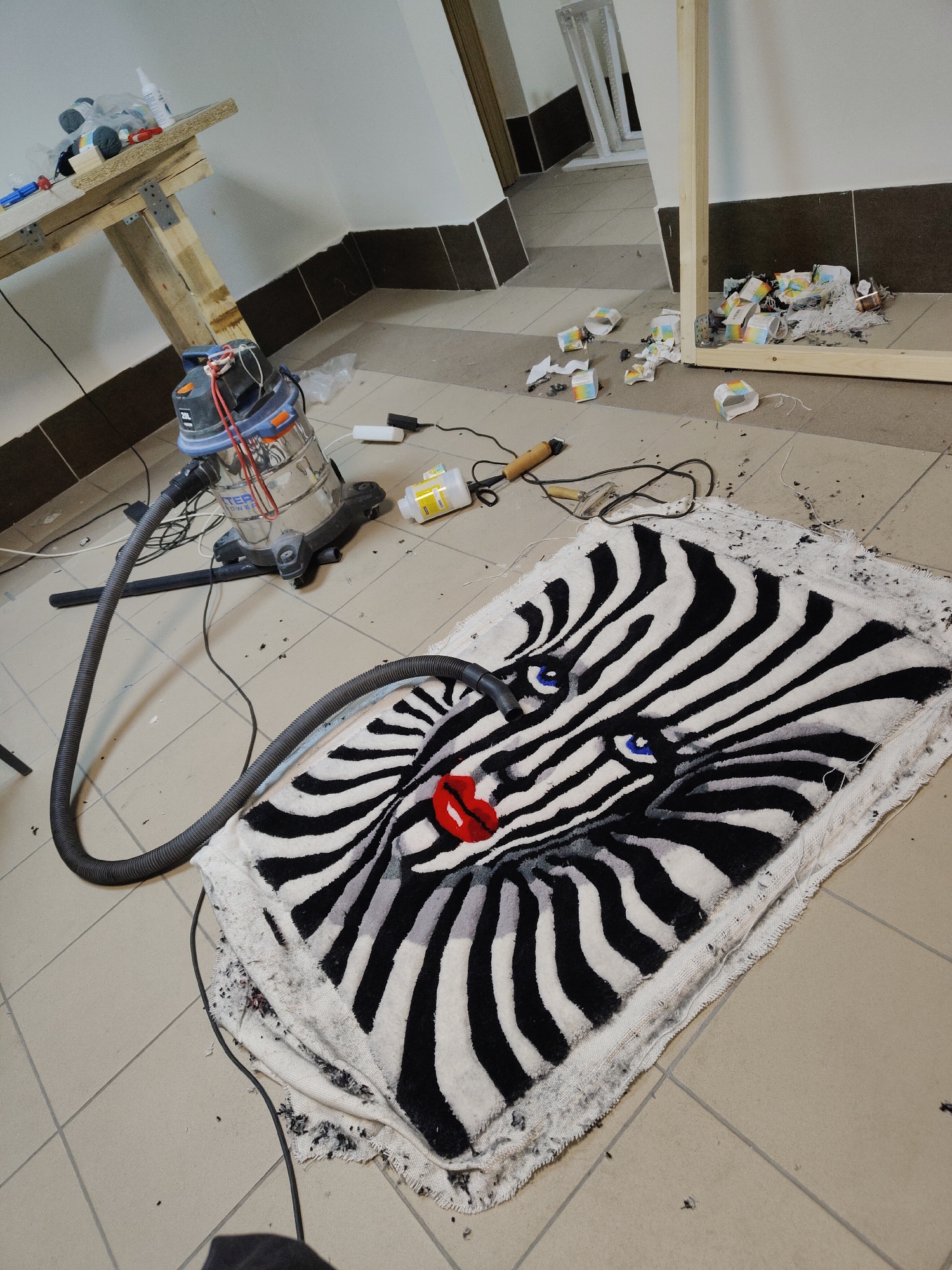 This media file depicts an indoor workspace dedicated to rug making, specifically illustrating the process of creating a tufted rug.
The central subject is a partially completed rug lying on a light-colored tiled floor. The rug features a bold black and white zebra-stripe pattern, forming the design of a face with vibrant blue eyes and striking red lips. The edges of the rug are unfinished and surrounded by numerous black and white yarn trimmings, indicating that work is still in progress. A vacuum cleaner hose is positioned near the rug, suggesting cleanup or trimming activity.
To the left of the rug, a wet/dry vacuum cleaner with a transparent top is visible. Text on its body reads "DEXTER POWER" and "20L". Its hose extends directly towards the rug. Further left, a rustic wooden table or workbench holds various small items including bottles and spools of thread.
On the right side of the frame, a large, upright wooden frame, likely used for stretching the rug backing during the tufting process, is present. Around its base and scattered on the floor are many empty, cylindrical yarn containers, some featuring a rainbow pattern, along with more yarn scraps.
Other tools and materials scattered on the tiled floor include a bottle with a yellow label (possibly glue), a heated tool with a power cord, and a small carving or trimming tool, all consistent with rug tufting equipment.
The setting is a functional room with light-colored floor tiles and plain white walls contrasted by a dark base trim. The scene is illuminated by indoor lighting. The visible debris and tools indicate active craftwork, with the vacuum cleaner ready for maintenance or finishing steps.