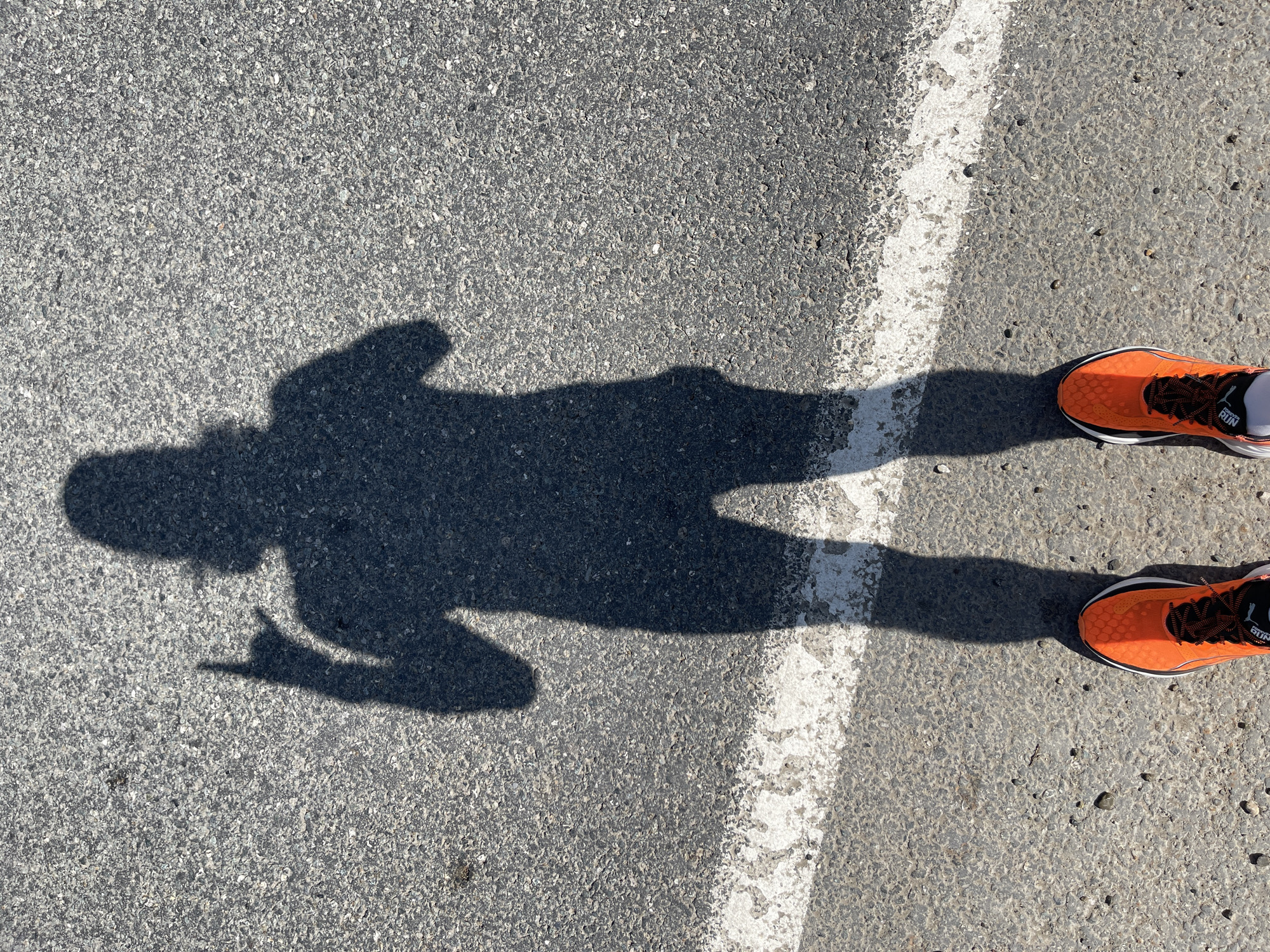 The image shows a ground-level view of asphalt pavement, partially bisected by a white painted line. A shadow of a person is cast on the pavement, oriented horizontally across the frame. The person appears to be standing, with one arm raised, perhaps holding something. The shadow suggests the person is wearing clothes or a dress. Adjacent to the right edge of the image are the bottoms of two feet wearing orange running shoes with black laces. The asphalt texture is visible, and the overall lighting suggests a sunny day. There is no way to know from the photo that the location is Перевозное, Russia.