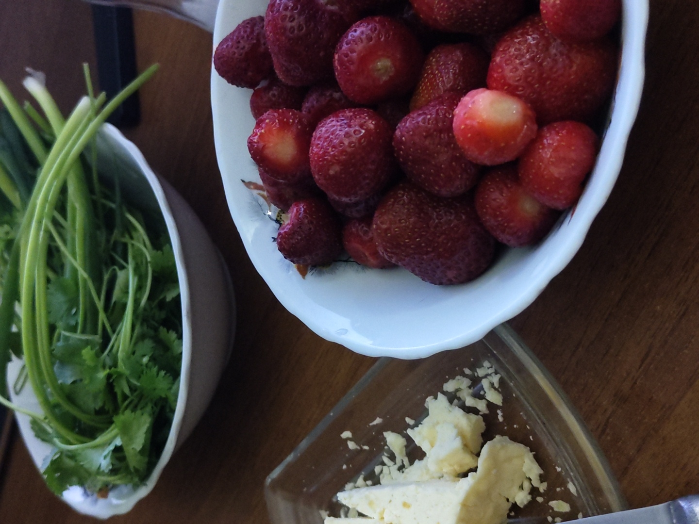 The image shows a close-up of a table with food items. In the foreground, a white bowl is filled with fresh strawberries. To the left, a clear glass dish contains chunks of white cheese, some of which are crumbled. In the background, another white bowl holds a bunch of green onions and cilantro. The table surface appears to be made of wood. The lighting suggests an indoor setting. There are no people or animals visible in the image.