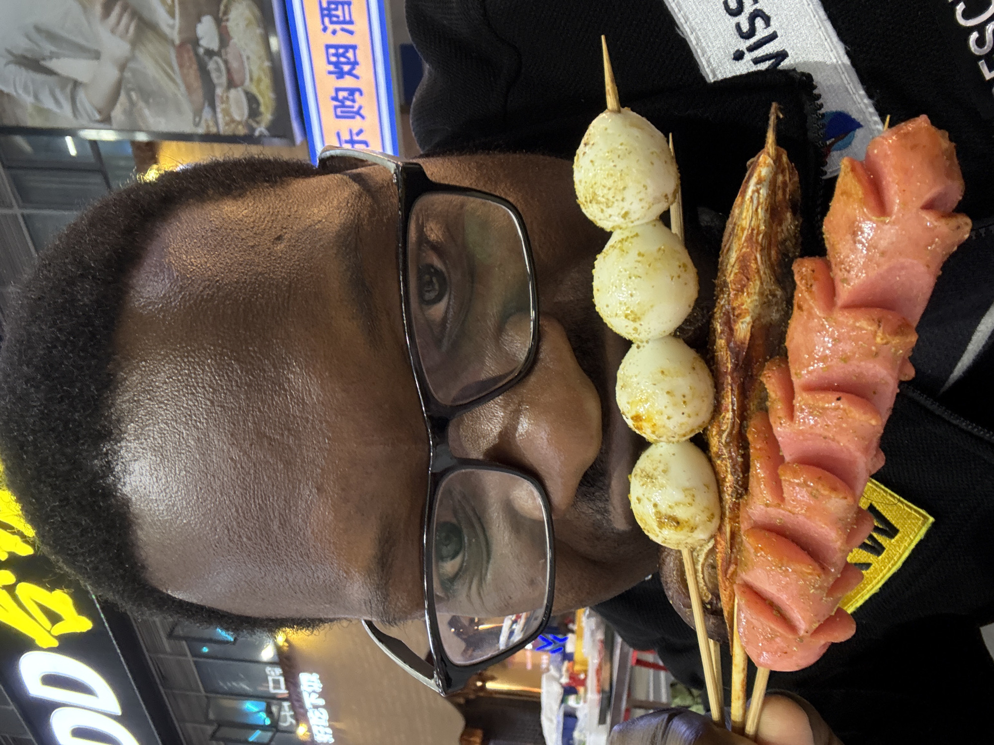 An adult male with dark skin and short, curly black hair, wearing black-rimmed glasses, is depicted in a close-up, eye-level shot, holding four skewers of food directly in front of his mouth. His mouth is partially open. The top skewer holds four round, white objects, possibly quail eggs, seasoned with golden-brown flakes. The second skewer contains a piece of grilled, seasoned fish. The third and fourth skewers feature pink, scored sausages, also seasoned. The man wears a black garment with a visible yellow embroidered patch and a white fabric label partially displaying "wiss". The background is a blurred, illuminated urban environment at night in Liangjiang, China, featuring various neon signs with Chinese characters. Visible signage includes "DD" and other characters, including "台" and "好吃不贵" (delicious and not expensive), along with another sign displaying "乐购烟酒" (Happy Shopping Tobacco and Alcohol) and an image of a person preparing food.