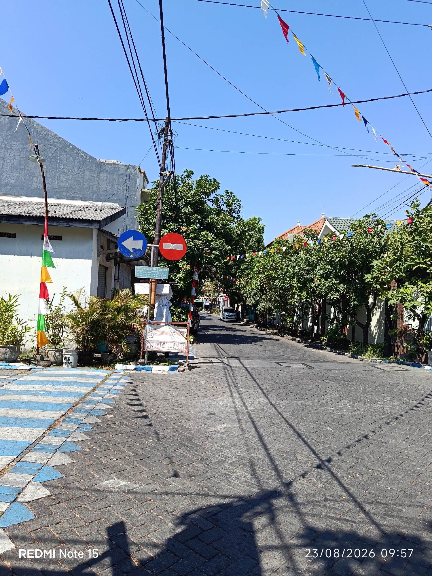 This is a photograph taken on a sunny day in the city of Keboansikep, Indonesia. The image depicts a street scene with a paved road and sidewalks. The road is made of dark gray interlocking pavers. The sidewalks have a decorative pattern of alternating blue and white painted stripes.
On the left side of the street, there is a building with a gray, textured wall and a white roof. In front of the building, several potted plants are visible. A flagpole with red, white, and green triangular flags is planted near the sidewalk. There is also a traffic sign with a blue arrow pointing to the left and another sign with a red circle and a diagonal bar, indicating "no entry" or "do not enter." A small, green sign with text is located below these traffic signs. Adjacent to the traffic signs is a white covered object on a stand.
On the right side of the street, a row of lush green trees lines the sidewalk, casting shadows on the road. Further down the street, houses with orange and green roofs are visible, interspersed with more trees. Colorful triangular flags are strung across the street overhead, adding a festive atmosphere. Electrical wires and poles are also prominent in the scene.
In the distance, a white car is parked on the right side of the road. The sky is clear and blue. The bottom left corner of the image displays "REDMI Note 15" and the bottom right corner shows the date "23/08/2026" and time "09:57".