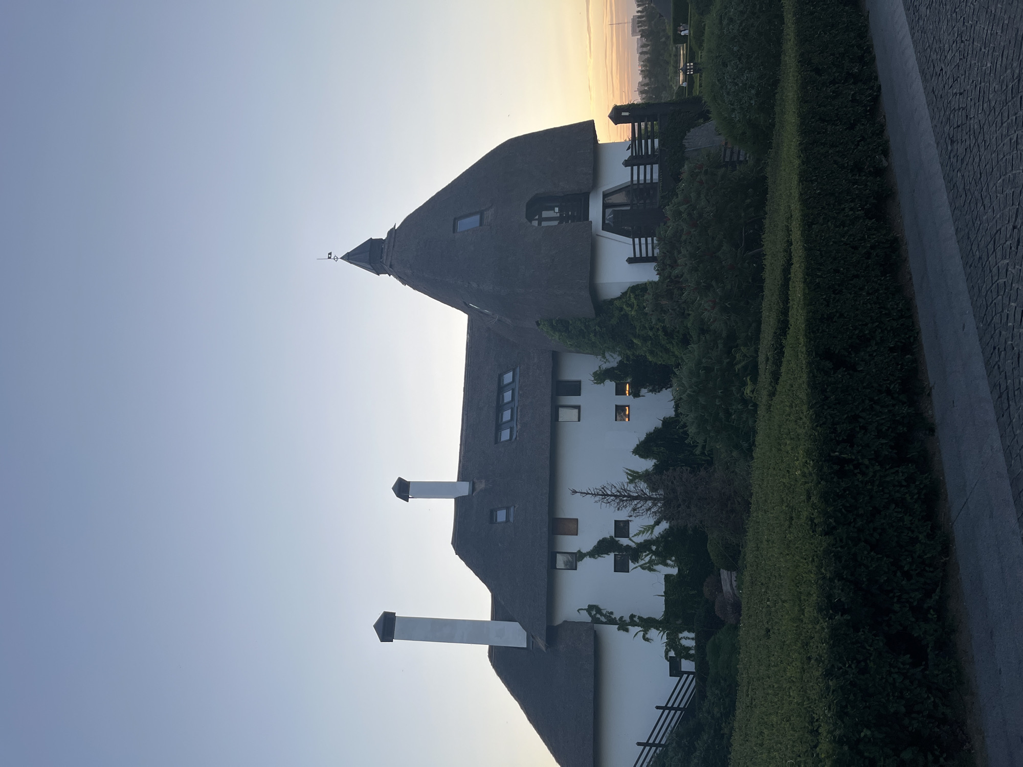 The image captures a large, white building with a thatched roof and two prominent chimneys, set against a twilight sky. The architecture features a steep, conical thatched section with a dormer window and a weather vane. The building is surrounded by lush green landscaping, including manicured hedges, trees, and climbing vines. A paved walkway leads in the foreground, and a wooden fence is visible along a sloped area to the left of the house. In the distance, a hazy skyline suggests an urban or suburban environment. The lighting indicates it is either early morning or late evening, with a warm glow on the horizon.
This building is located at the address Soborna Square, 11, in the city of Dnipro, Ukraine.