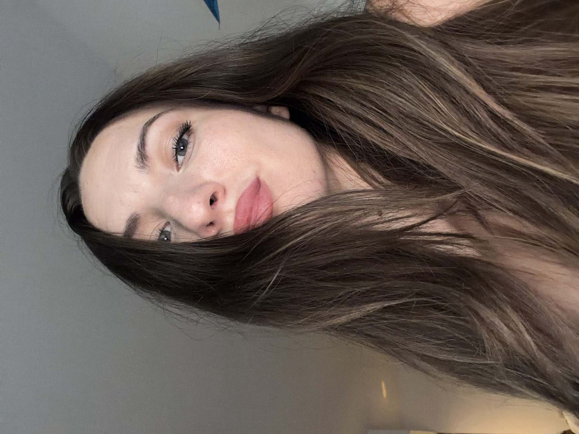 The image is a close-up selfie of a woman with long, brown hair. Her face is turned slightly to the side, and she is looking at the camera with her lips pursed. She is wearing makeup, and her hair is partially obscuring her face. The background appears to be an indoor setting with a gray wall. There is no identifiable information within the image to suggest a location in Sharm El Sheikh, Egypt.